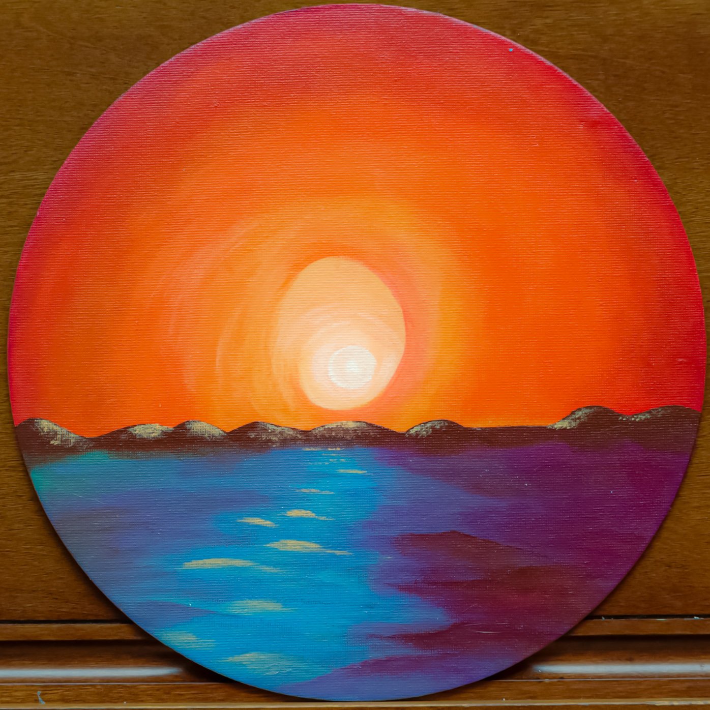 A circular painting of a sunset over mountains and water, with an orange and pink sky, snow-capped mountains, and reflections in the blue water.