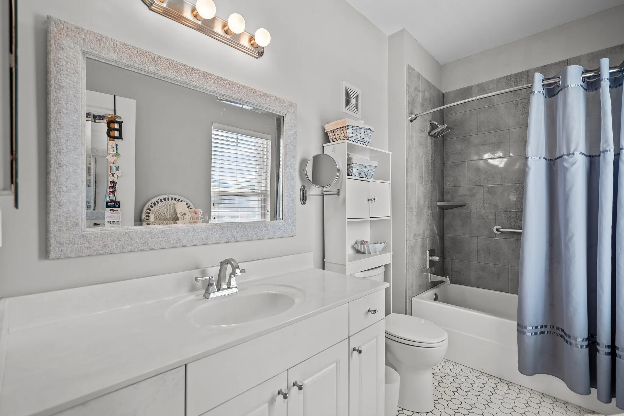 Bathroom with white vanity mirror, sink, faucet, window, and shower with gray tiles and a blue shower curtain.
