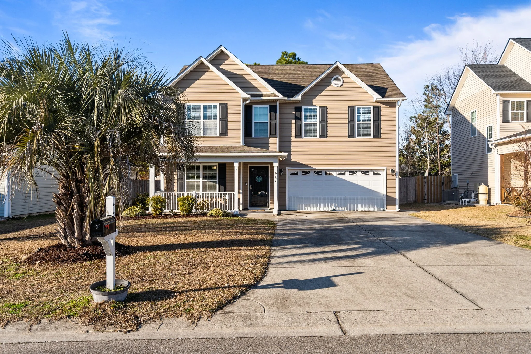 Front view of a two-story suburban house with beige siding, black shutters, a white garage door, and a small front porch with white railing. There is a mailbox on a post next to a palm tree in the yard. The driveway leads up to the garage, and neighboring houses are visible on either side. The sky is clear and blue.