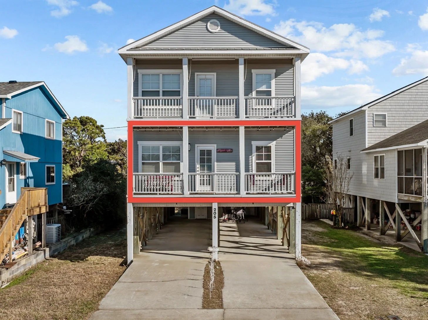 A three-story beach house on stilts with gray siding, white trim, and a balcony on each level. The middle balcony is outlined in red. The house has a concrete driveway leading to a parking area underneath.