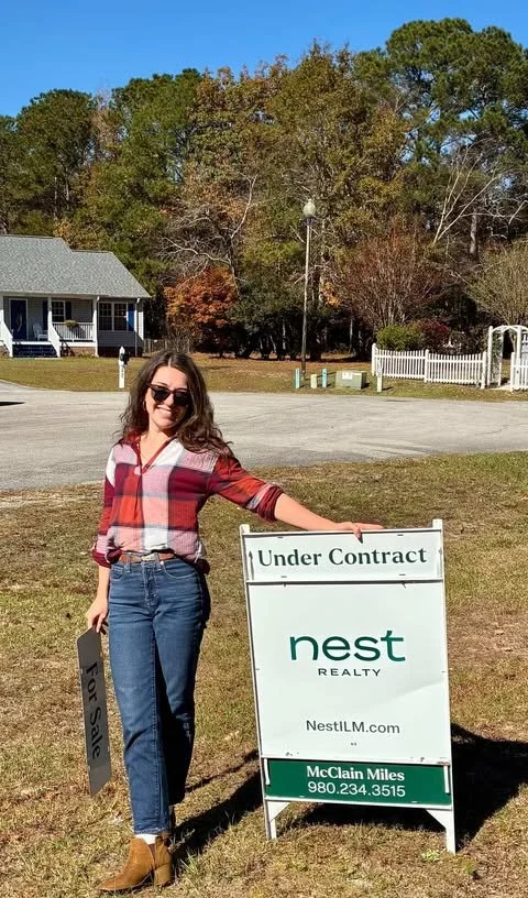 A woman with sunglasses and long hair, wearing a plaid shirt and jeans, standing outdoors near a sign that says 'Under Contract' and 'Nest Realty,' with a house and trees in the background.
