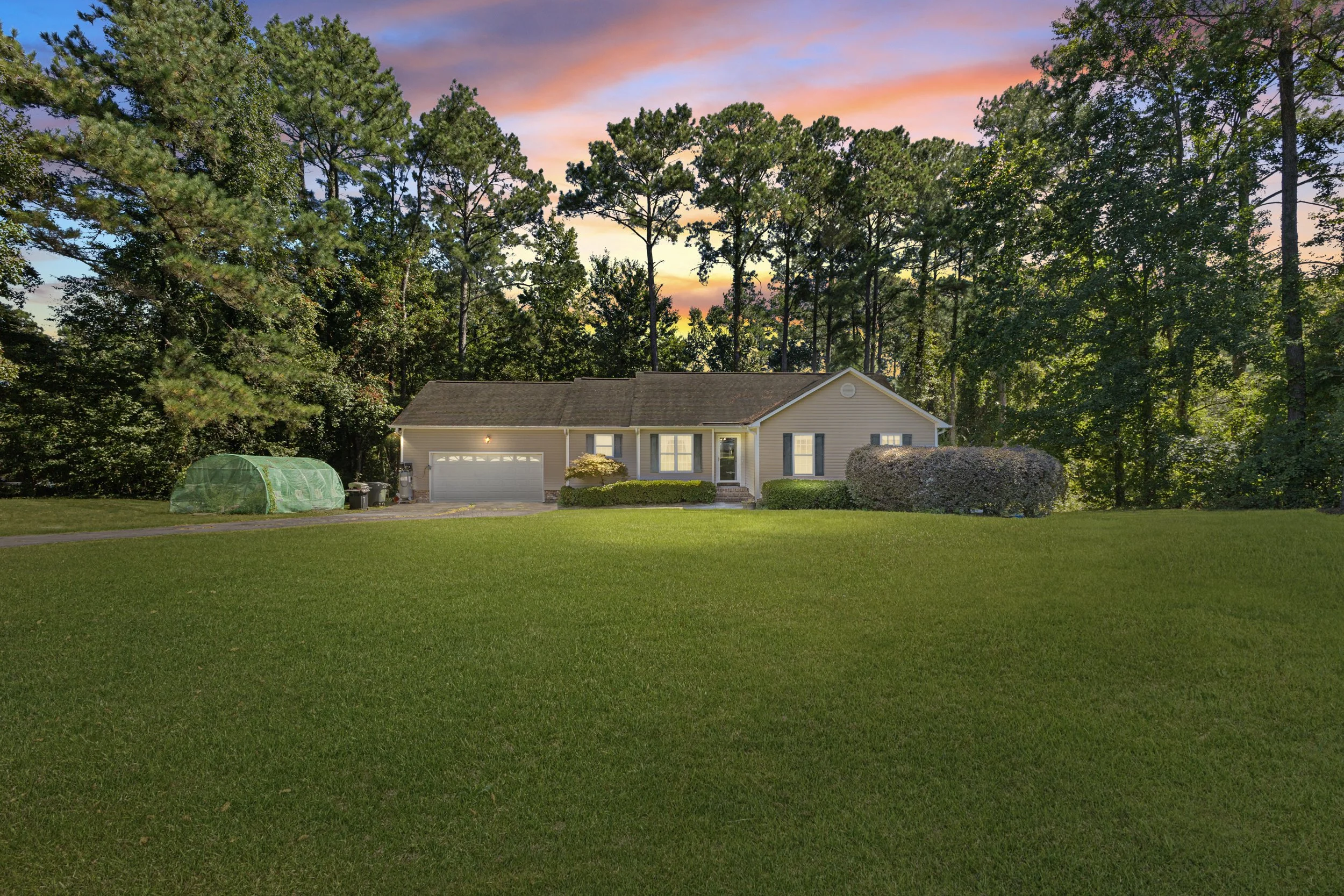 A single-story house with a two-car garage, surrounded by a green lawn and trees, during sunset