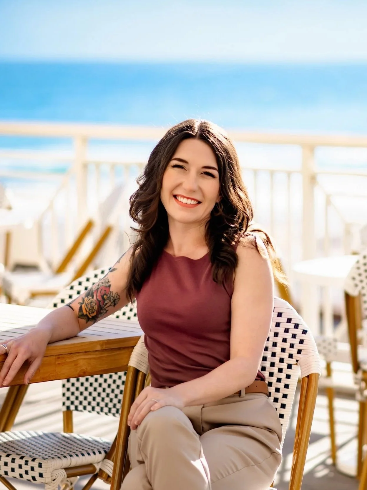 A woman with dark wavy hair, wearing a sleeveless maroon top and beige pants, smiling while seated outdoors near the water, with a tattoo of flowers on her left arm.