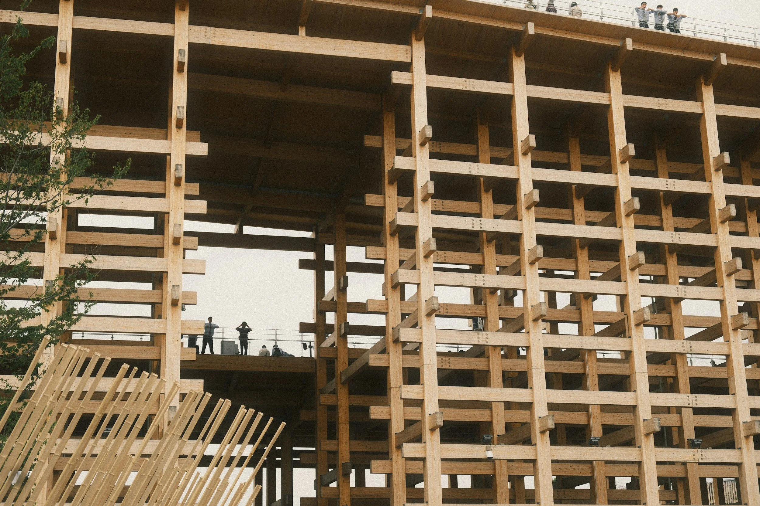 The image shows a large wooden structure under construction, with multiple vertical and horizontal beams creating a grid pattern. There are some people on the upper level, observing or working on the site. A tree is partially visible on the left side.