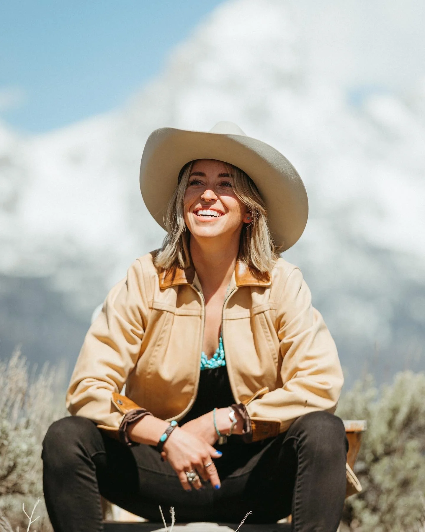 Woman sitting outdoors, smiling, wearing a large beige hat, a tan jacket, and turquoise jewelry, with cloudy sky in background.