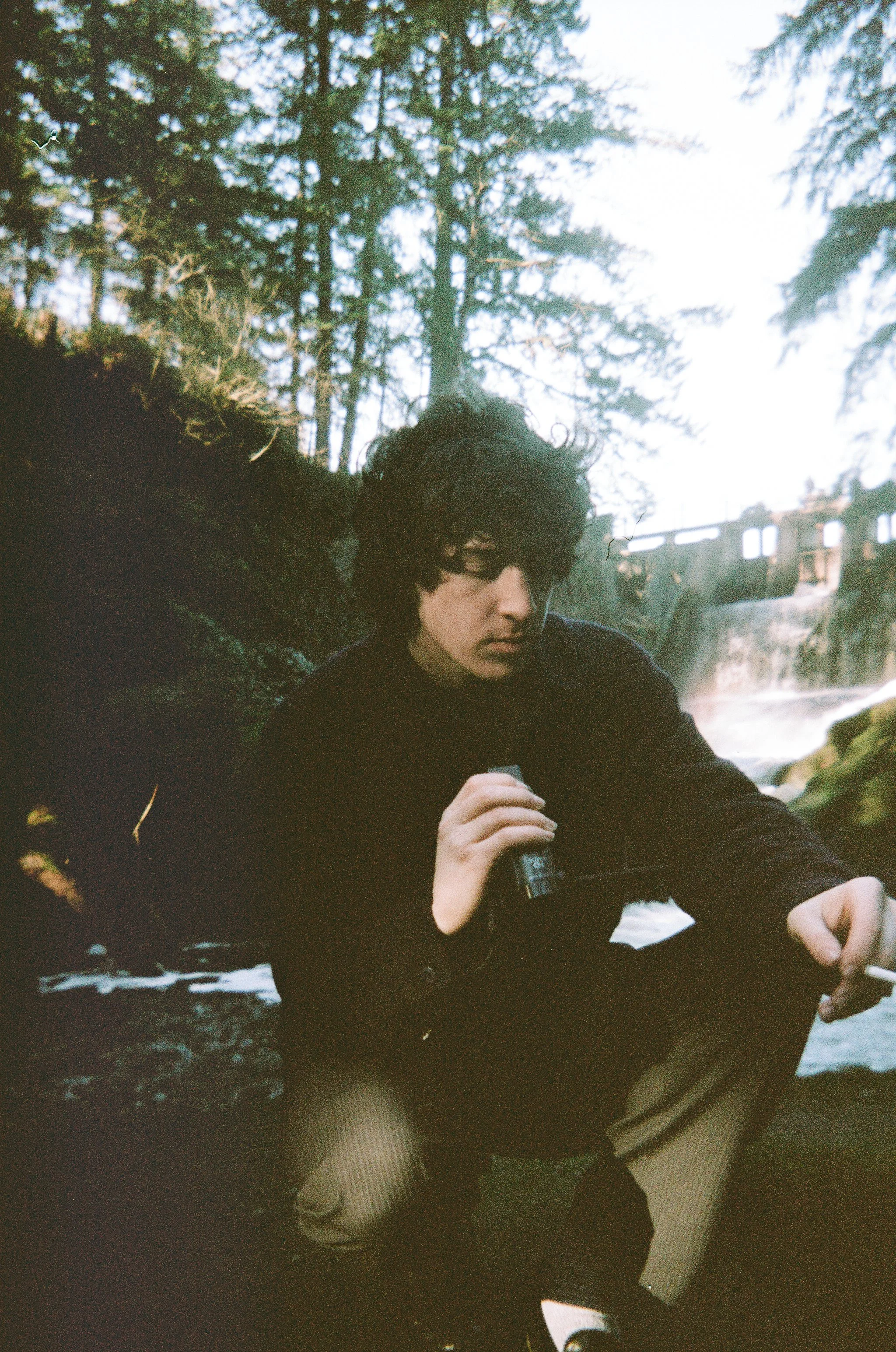 A young man with dark hair and glasses sitting outdoors by a river, holding a small object in his right hand and a cigarette in his left, with a backdrop of trees and a small waterfall.