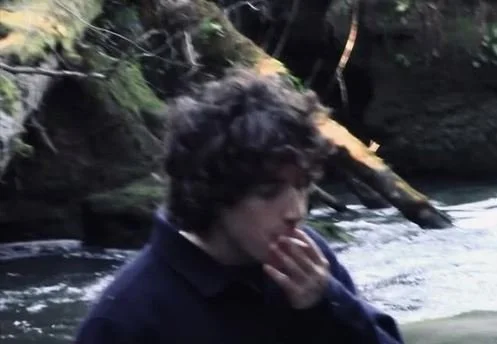 Person with dark curly hair wearing a dark jacket near a river or stream with rocks and trees.