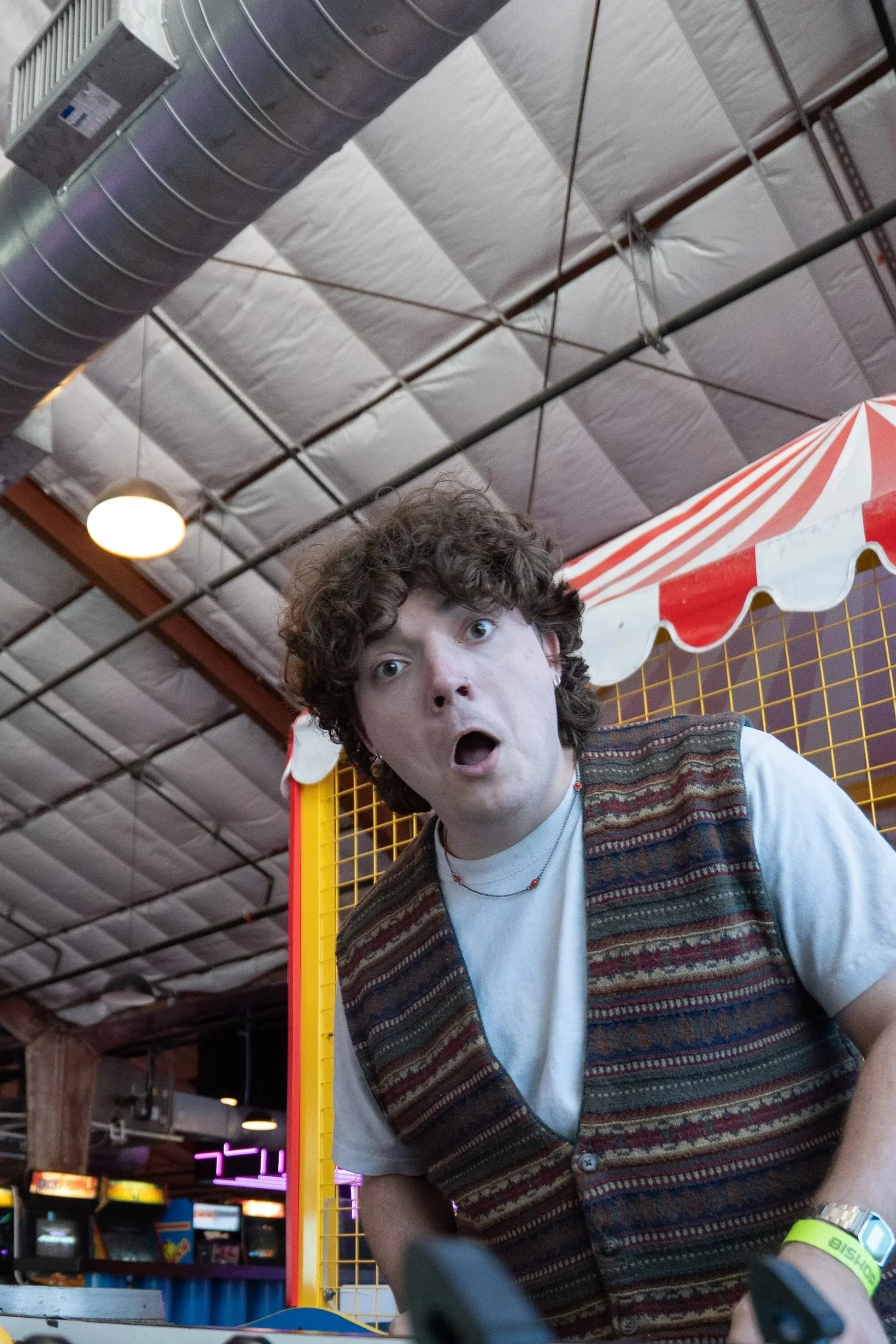 A young man with curly hair and surprised or shocked expression at an indoor arcade or entertainment center. He is wearing a colorful patterned vest over a white t-shirt and has a yellow wristband.