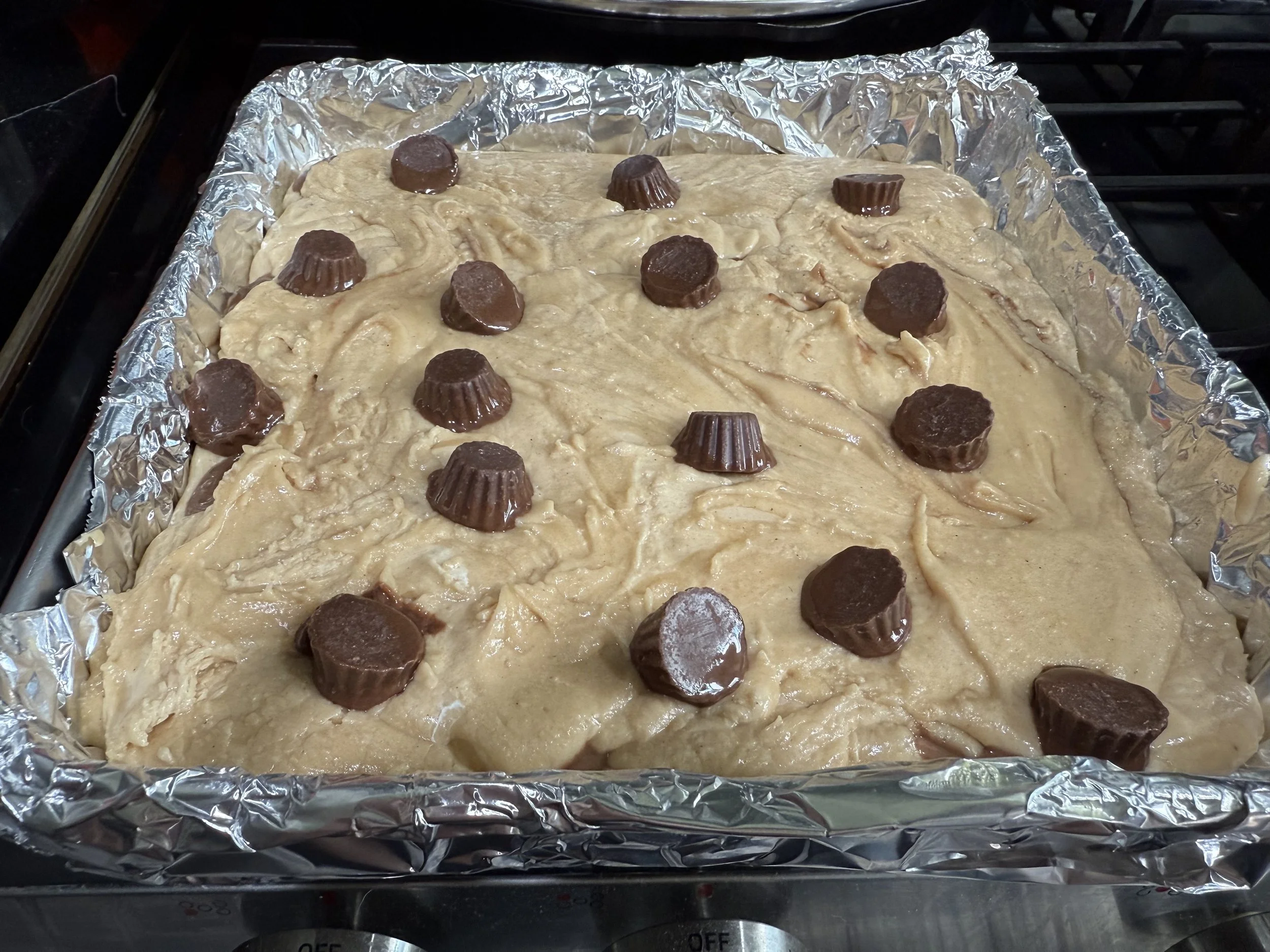 Unbaked blond cookie dough with Reese's peanut butter cups placed on top, in a baking pan lined with aluminum foil.