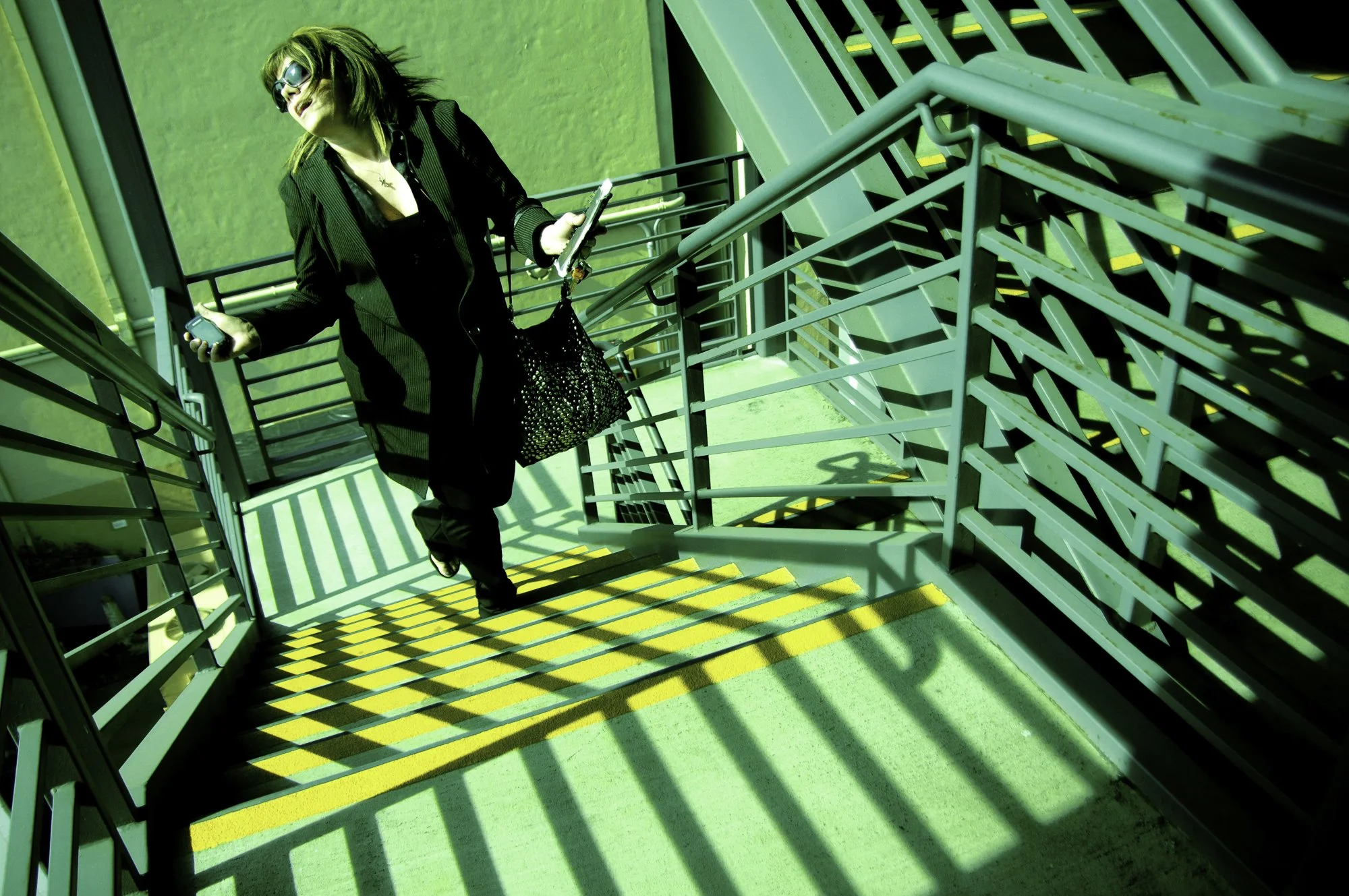 Woman with sunglasses, black jacket, and black pants walking down a neon green staircase with yellow safety markings, holding a phone in one hand and a bag in the other.