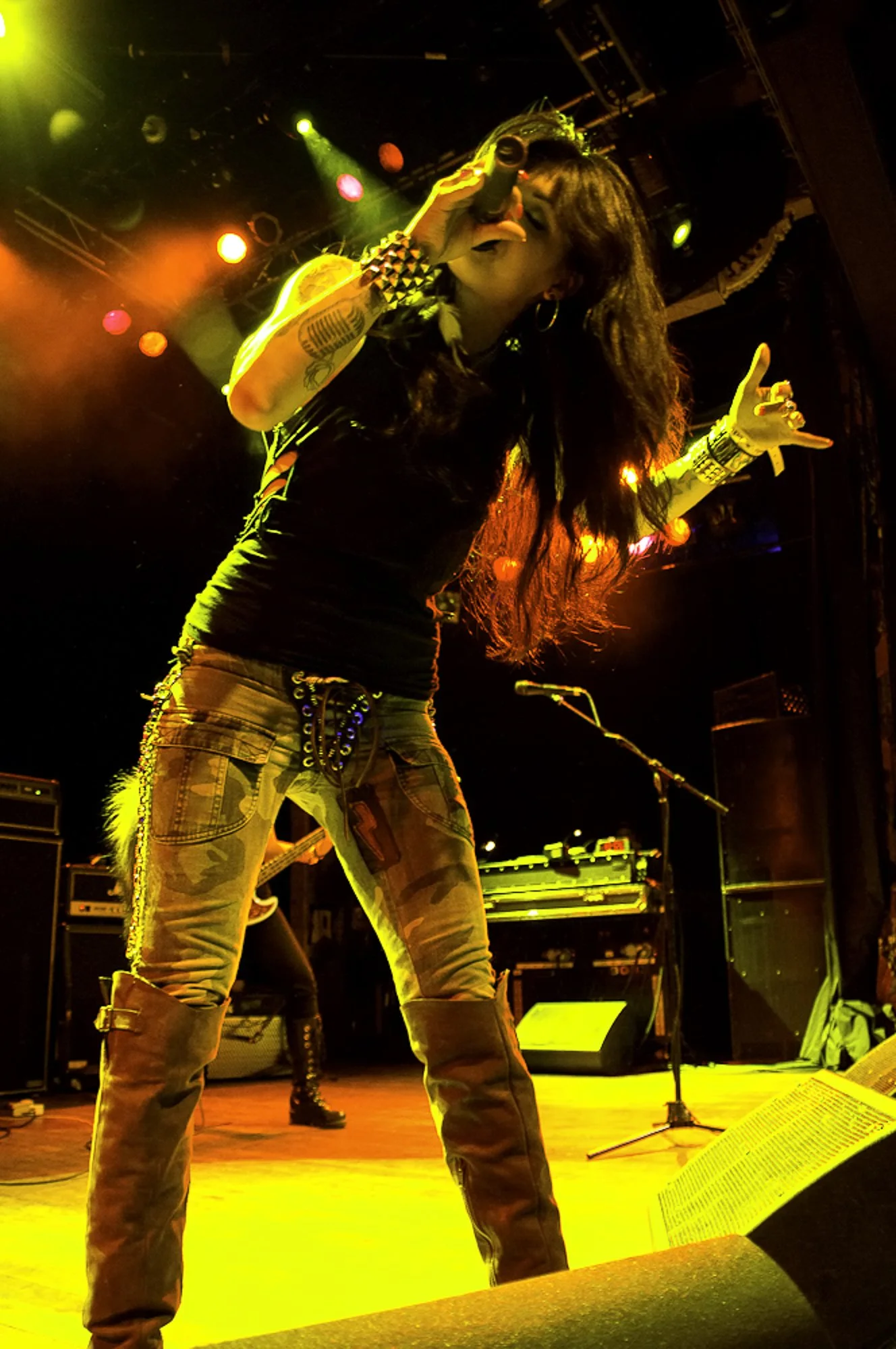 Female singer performing on stage with black shirt, camouflage pants, and long dark hair, holding a microphone and making a rock gesture, under colorful stage lighting.