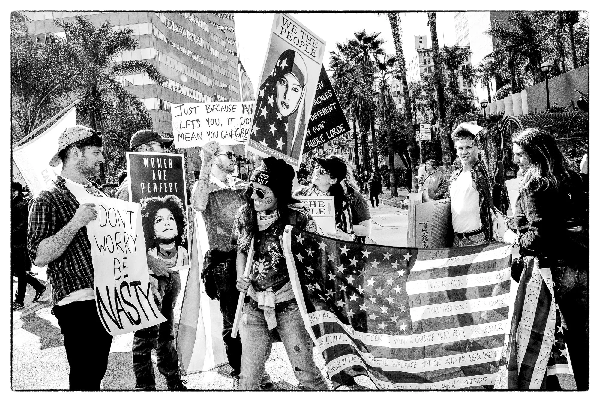 People participating in a protest or rally on a city street, holding signs and banners advocating for women's rights and social justice. Palm trees and tall buildings are visible in the background.