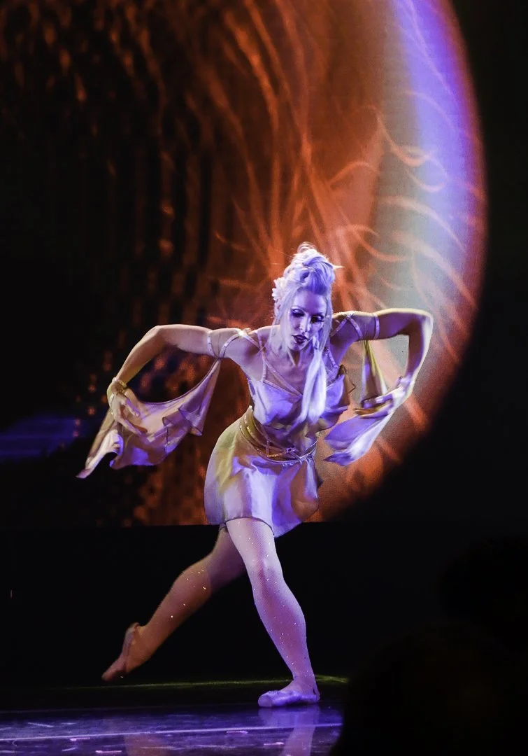 Performer with white hair dressed in a flowing costume dancing on stage with a colorful background projection.