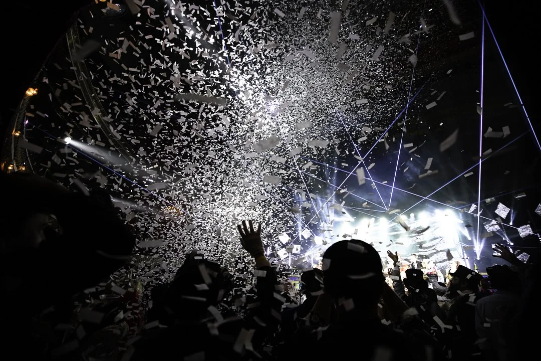 Concert or event scene with confetti falling, people raising hands, bright stage lighting, and blue laser lights.