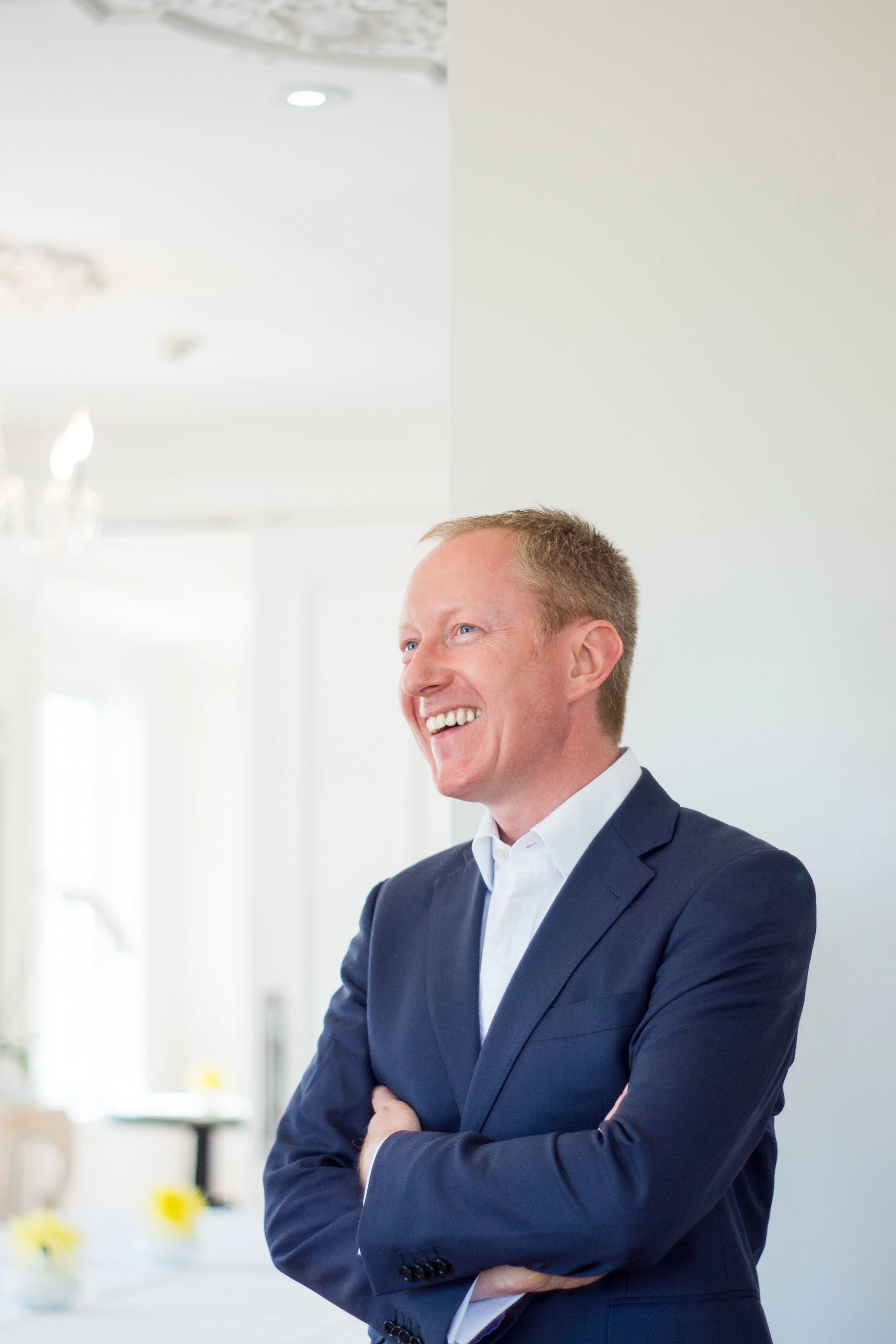 A man in a navy suit smiling with his arms crossed in a bright, elegant room.