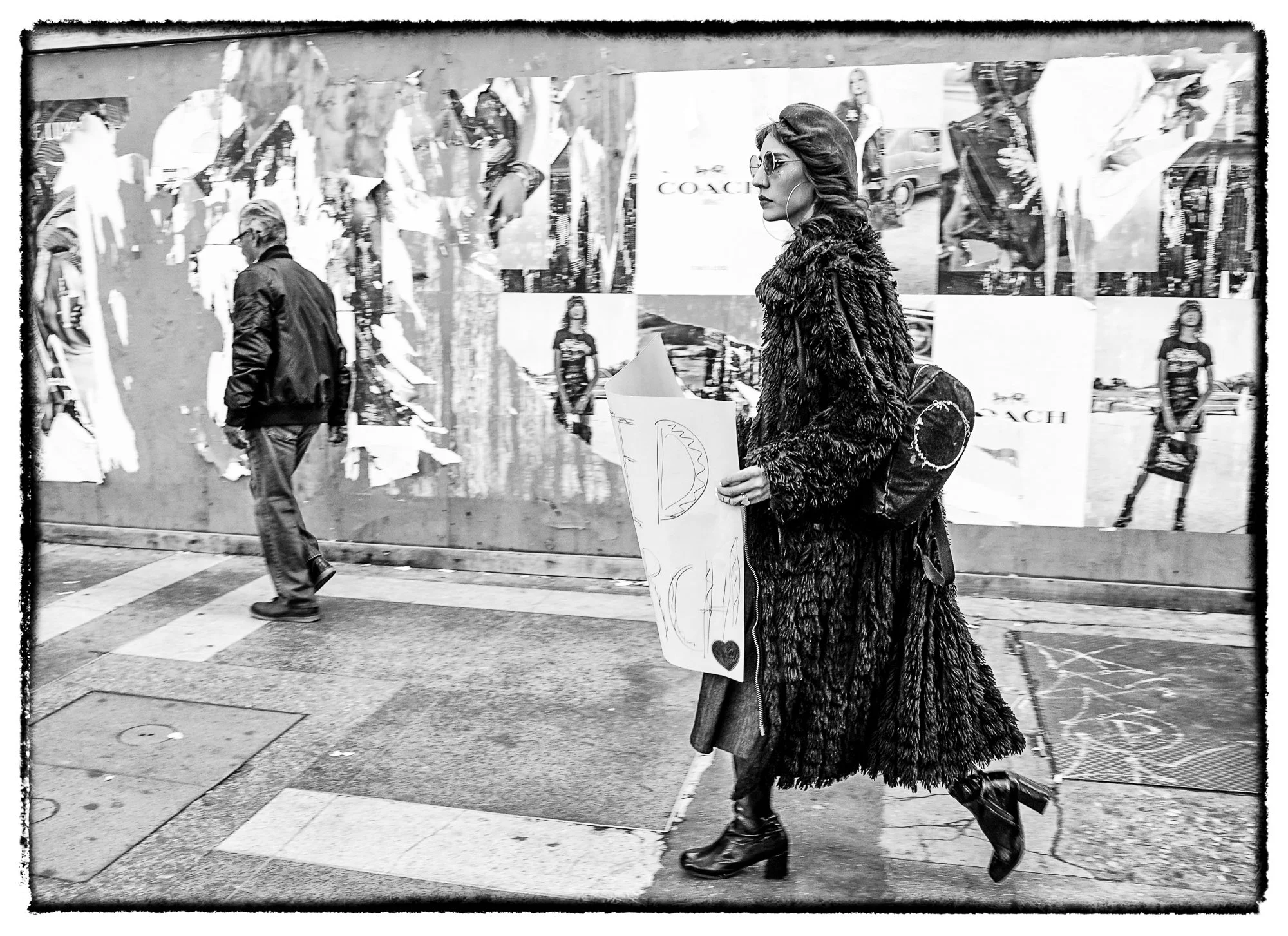 A woman walking on a city sidewalk holding a sign with a drawing and a heart, wearing a fur coat, sunglasses, and heeled boots, with a backpack. A man is walking past in the background, and there are posters and advertisements on the wall behind them