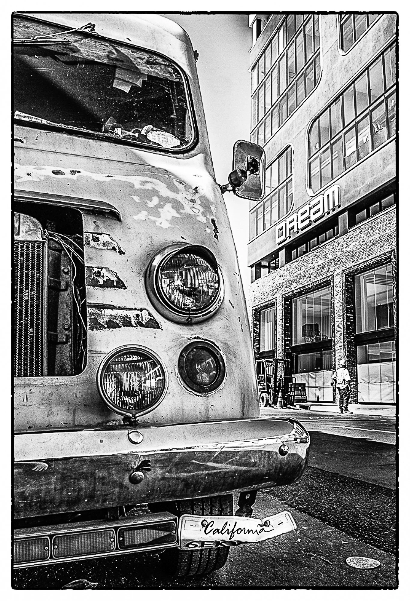 Black and white photo of an old, weathered bus with a California license plate, parked on a city street. Modern buildings and a pedestrian walking in the background.