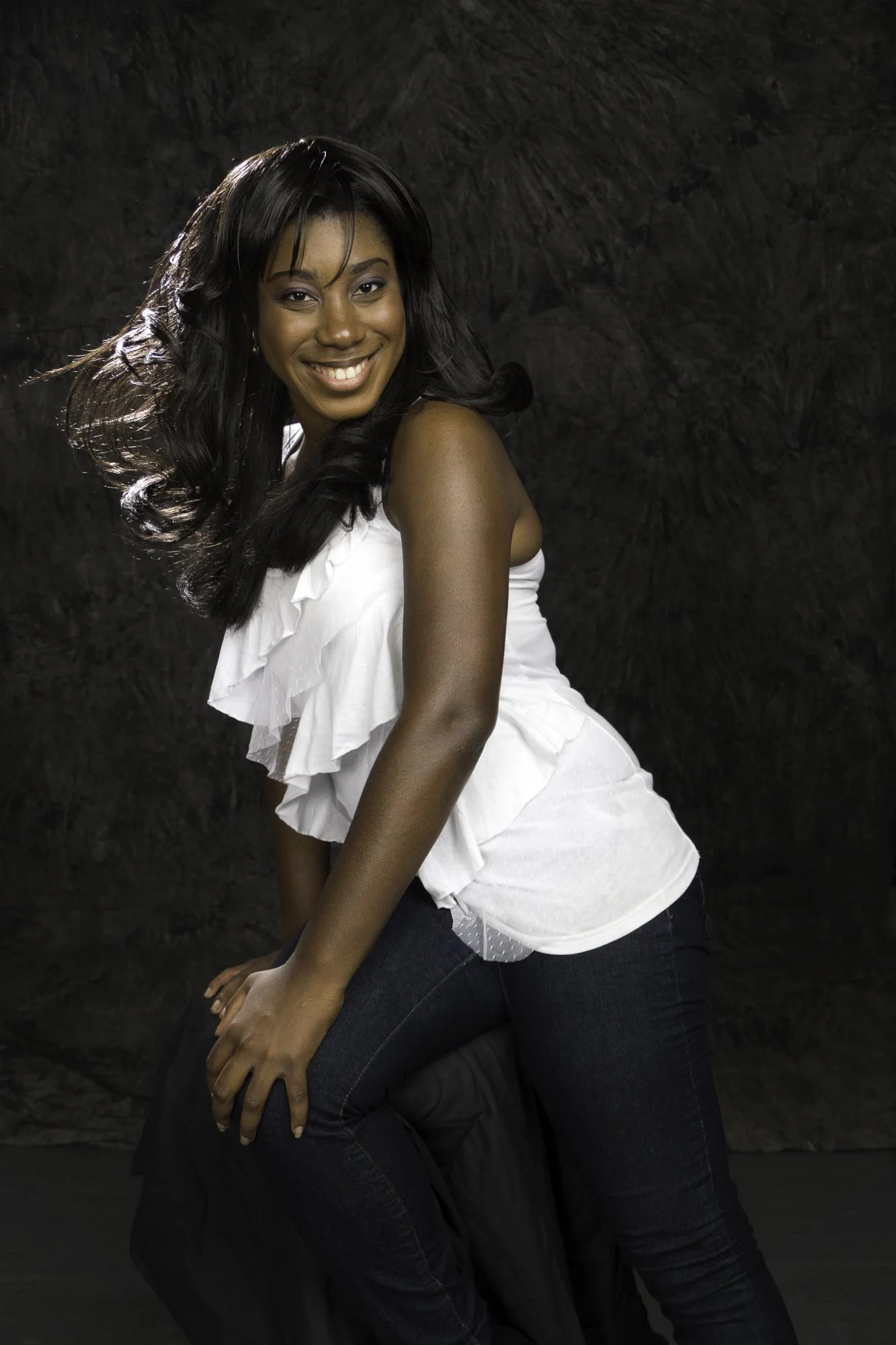 Portrait of an African-American woman with long black hair and a white ruffled top smiling and posing against a dark brown textured background.