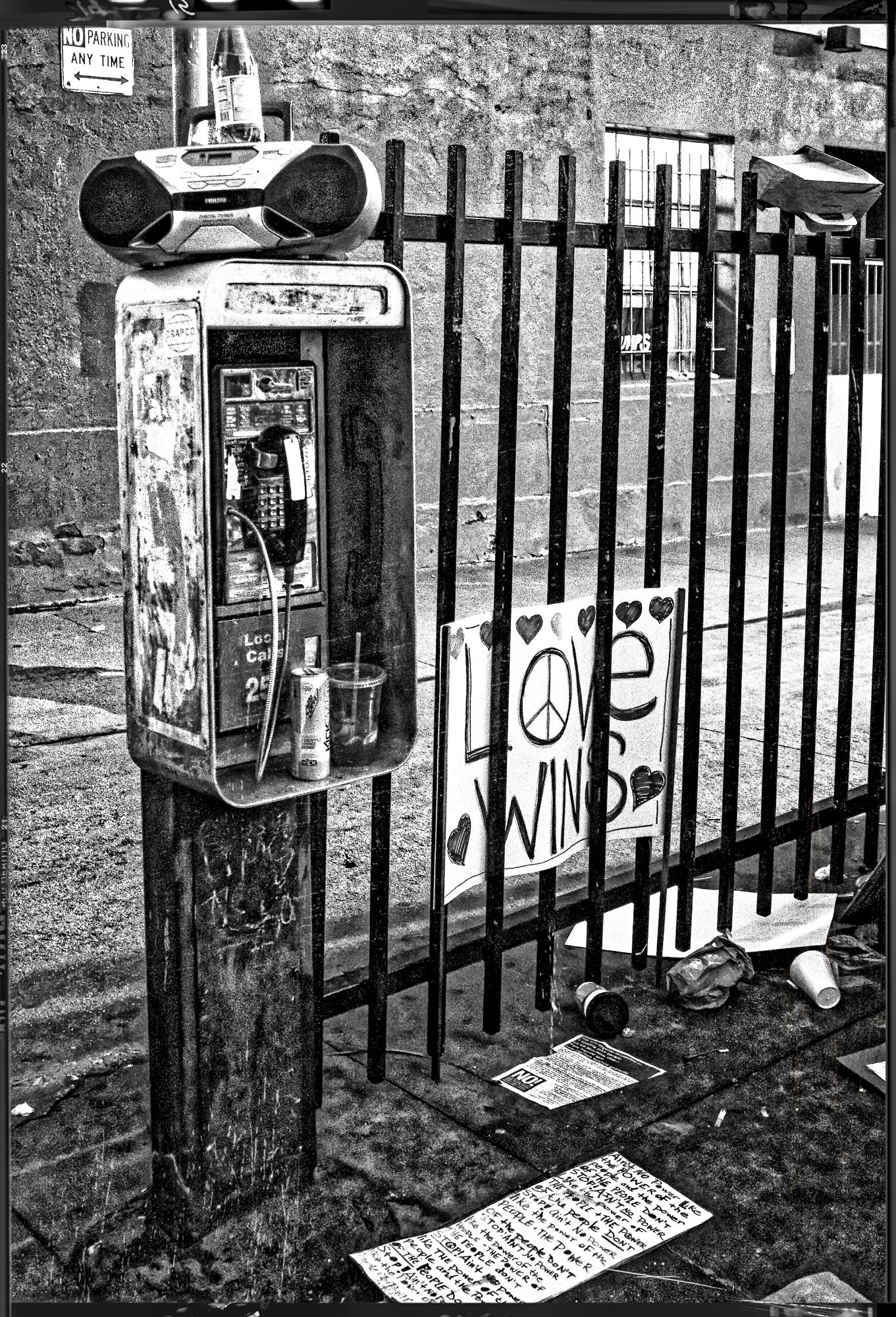 An old payphone booth with a boombox on top, a sign that says 'LOVE WINS' with peace symbols and hearts, and littered trash on the ground.