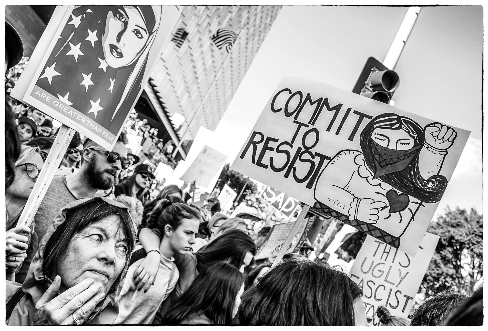 Crowd of protesters at a rally holding various signs, including one that reads 'Commit to Resist' with a drawing of a woman wearing a mask and raising her fist.
