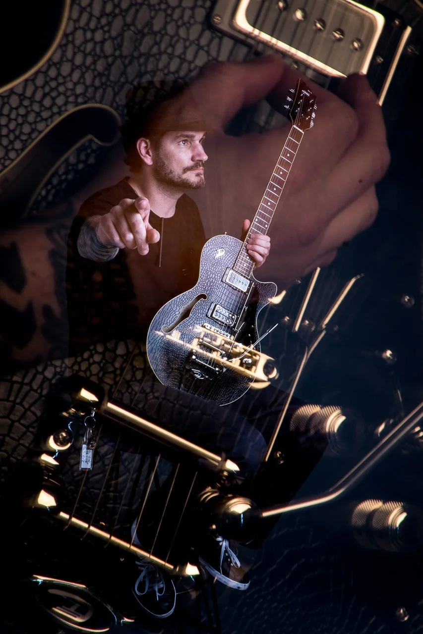 Reflection of a man with a guitar, pointing, surrounded by musical equipment and a textured surface.
