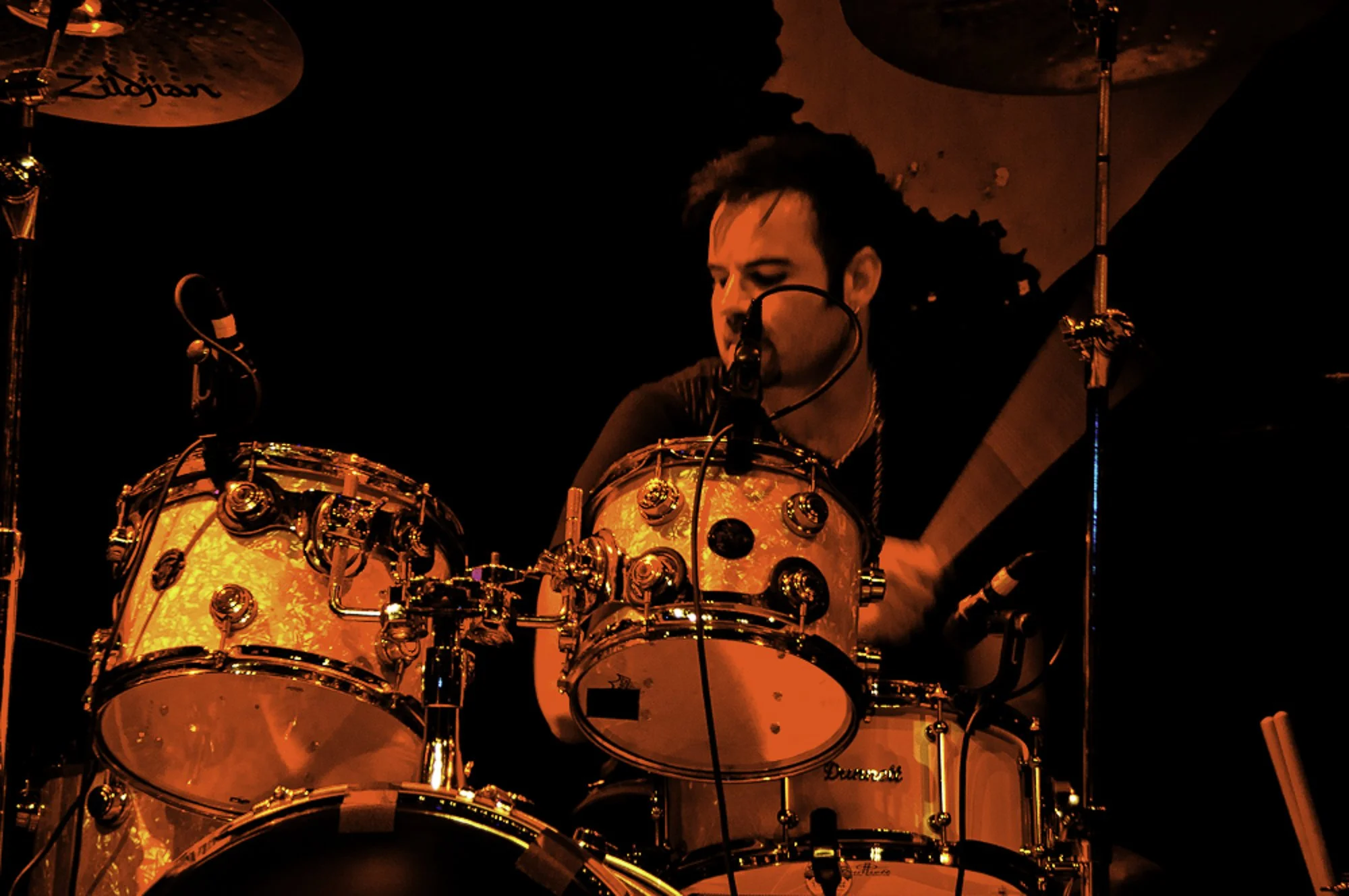 A drummer playing drums on stage, illuminated by red lighting, with a dark background.