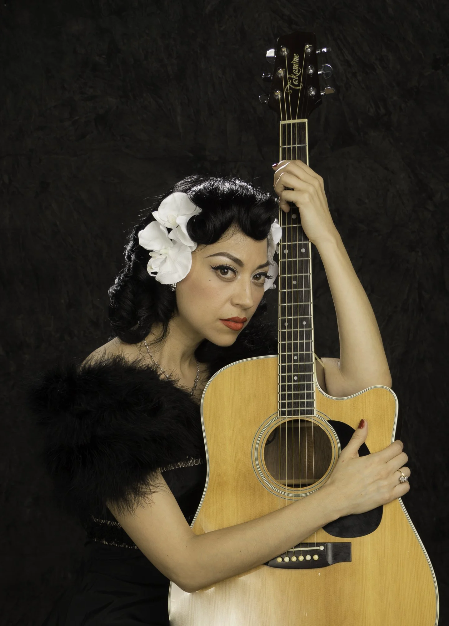 Portrait of a Mexican-American woman with vintage style hair and makeup, wearing a black dress with a fur shawl and flowers in her hair, holding an acoustic guitar.
