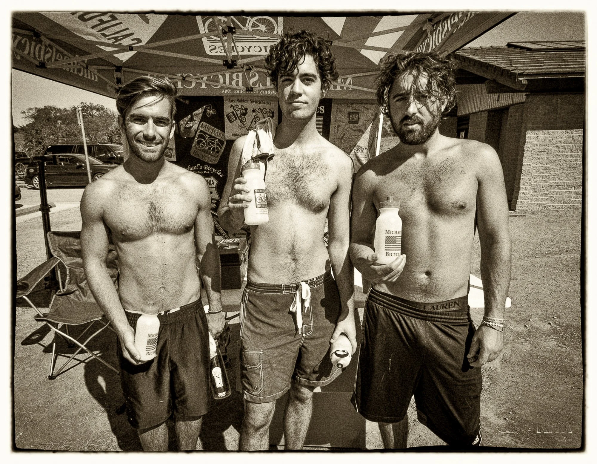 Three shirtless young men standing outdoors, each holding a bottle of sunscreen lotion. They are under a canopy with banners promoting bicycles. The background shows parked cars and a building.
