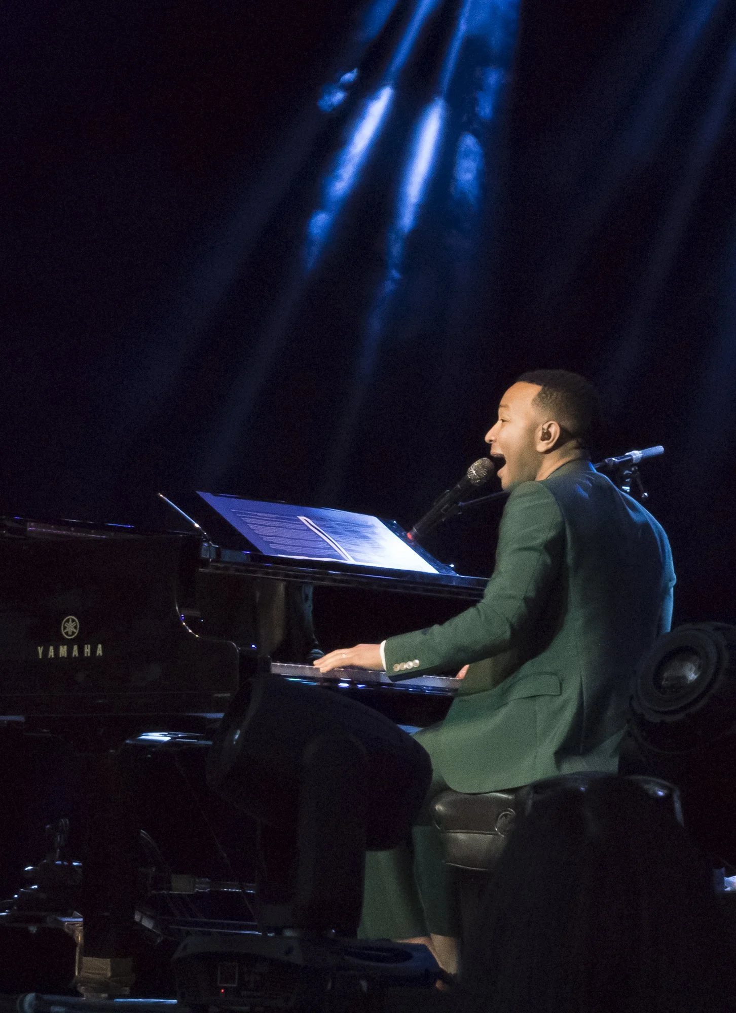 A man in a green suit playing a Yamaha piano and singing into a microphone on a dark stage with blue stage lights.