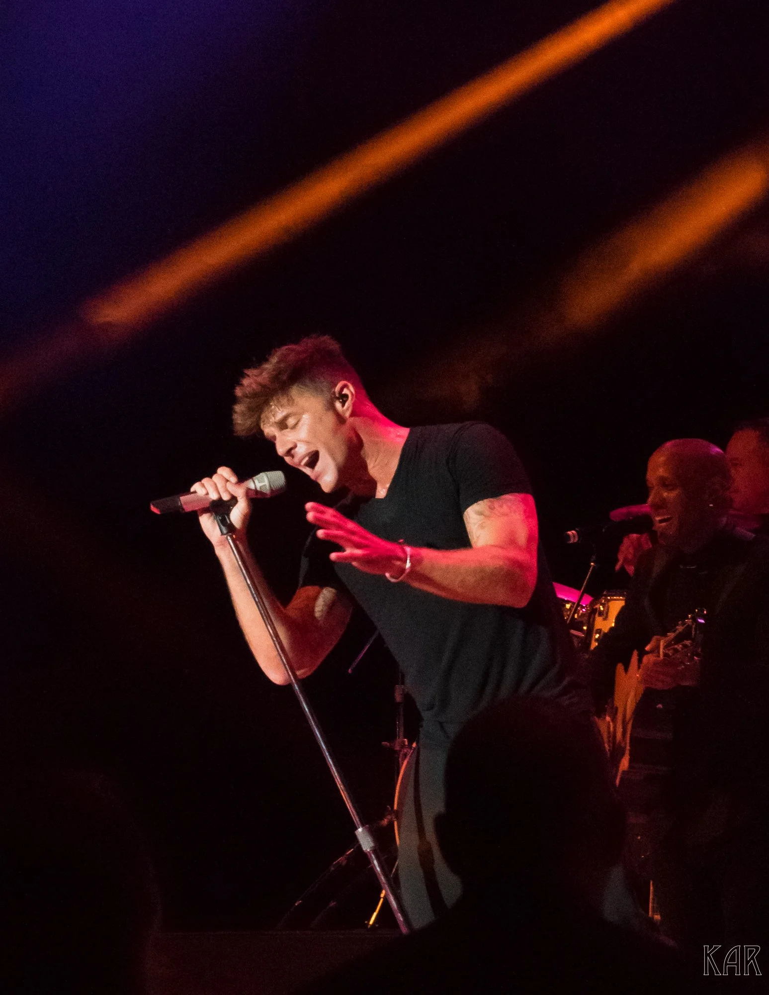 A male singer passionately performs on stage holding a microphone, with backup musicians smiling behind him, under colorful stage lighting.