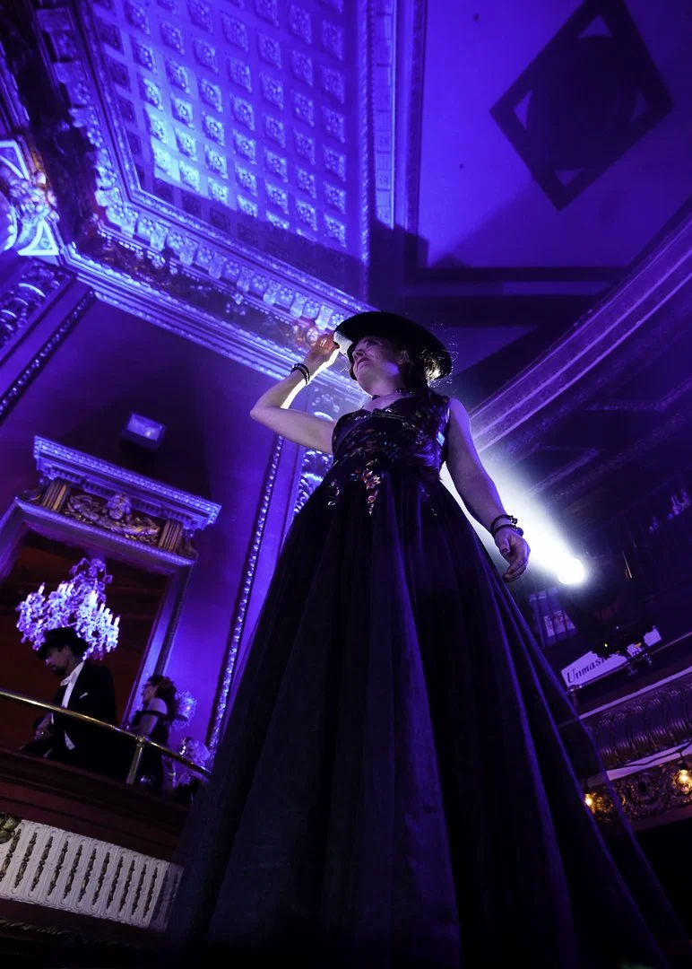 A woman in a long black dress and a wide-brimmed black hat standing on a stage with dramatic blue and purple lighting in an ornate theater or opera house.