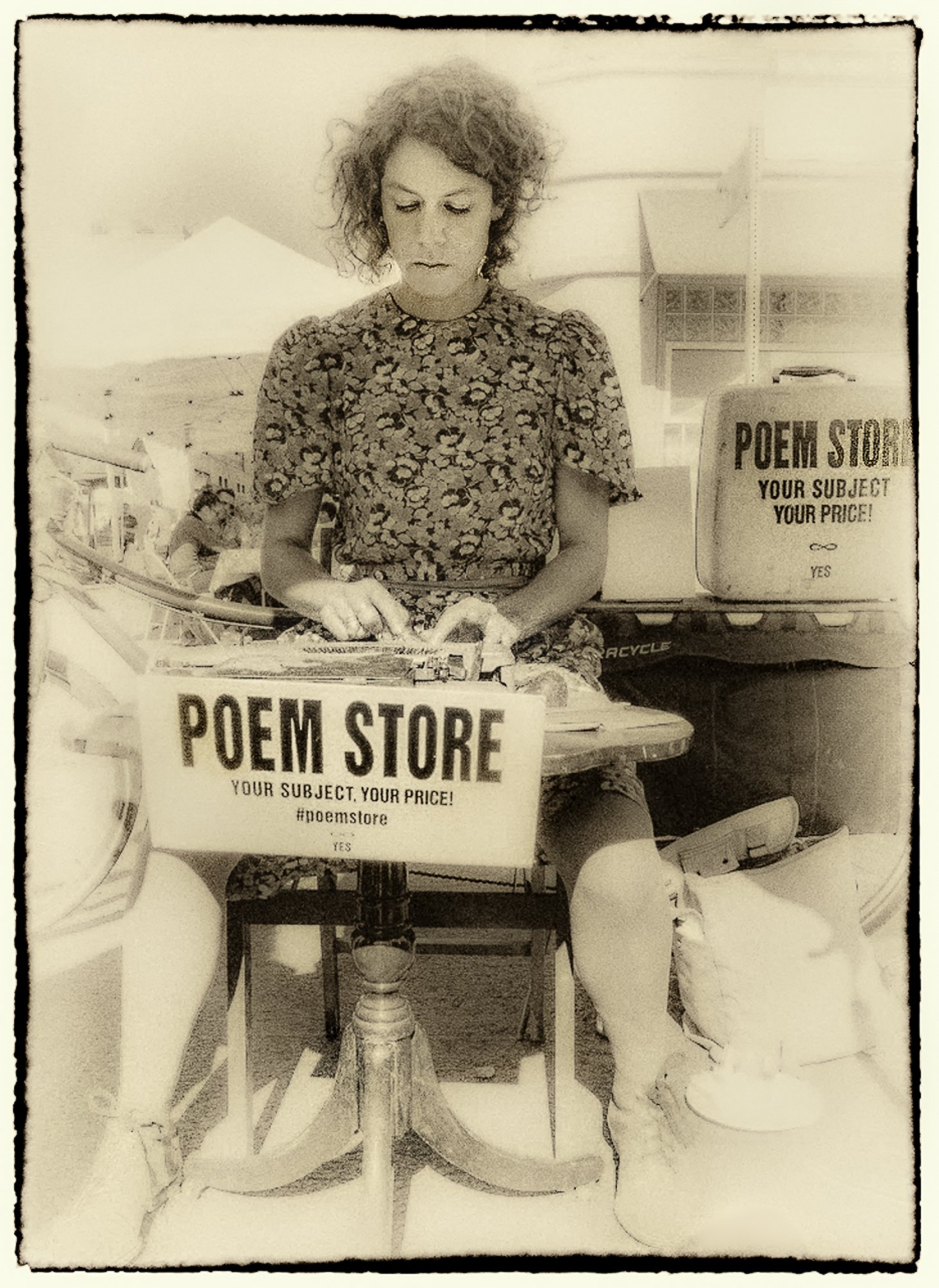 A woman with curly hair sitting at a table with signs that read 'Poem Store, Your Subject, Your Price! #poemstore'. She appears to be writing or reading something in front of her.