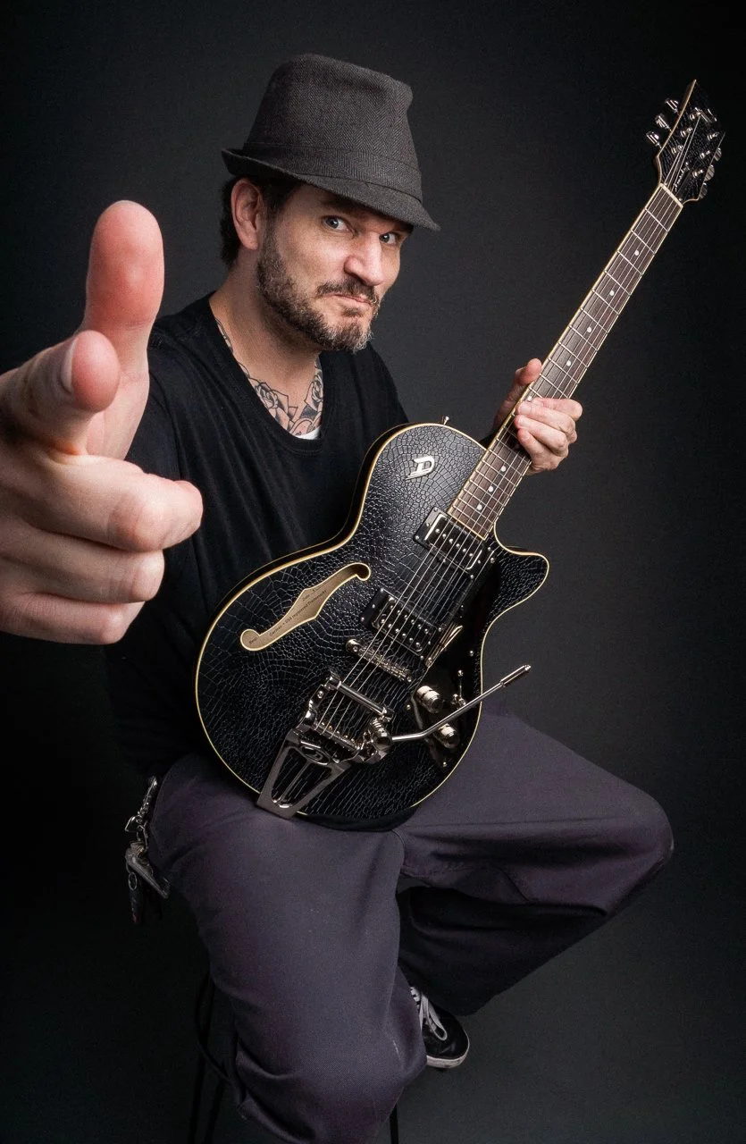 A man with a dark hat and black shirt holding a black electric guitar, sitting against a black background, pointing towards the camera with a serious expression.