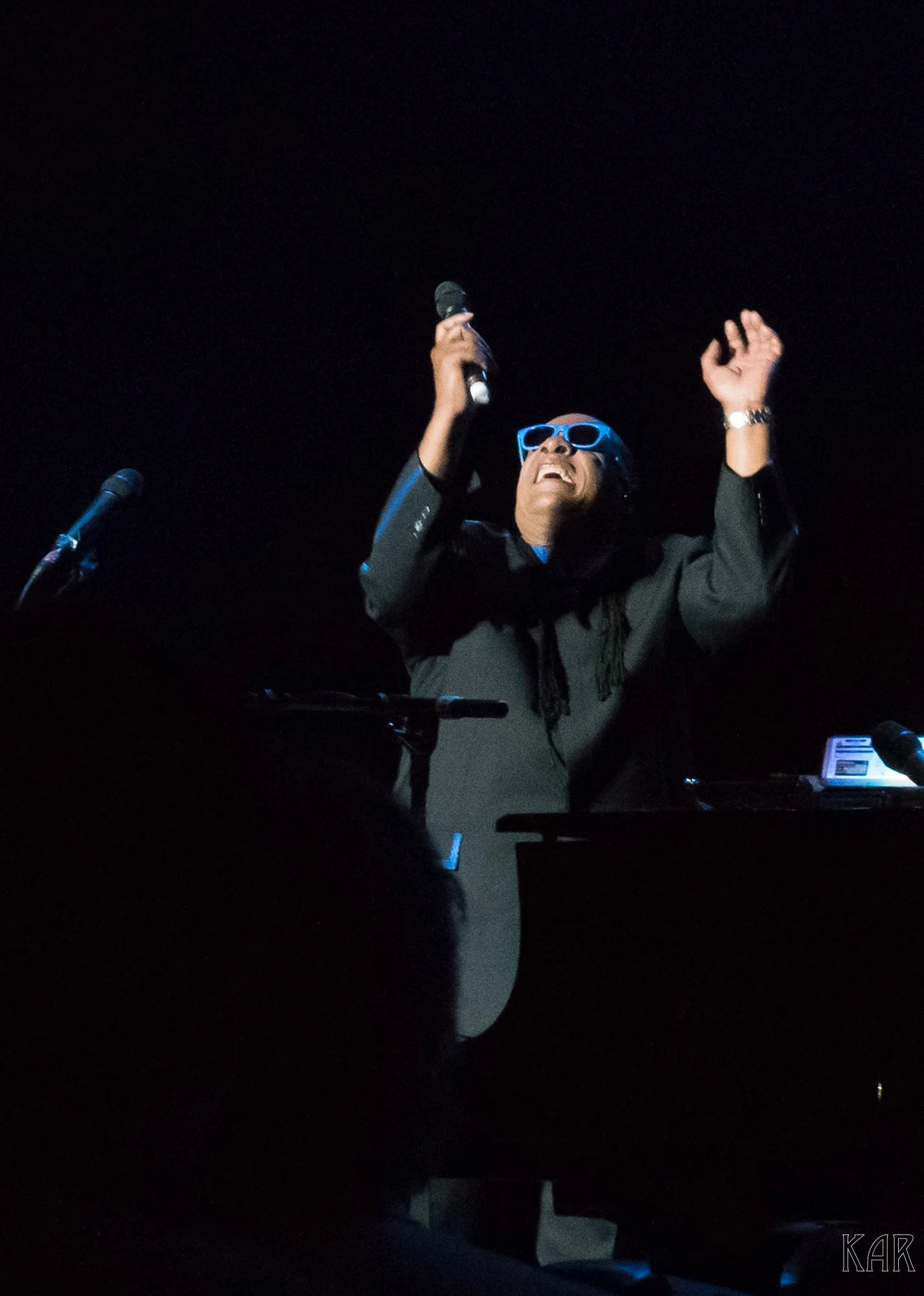A man with dark hair and glasses holding a microphone, singing with his eyes closed and head tilted back, standing behind a keyboard on a dark stage.