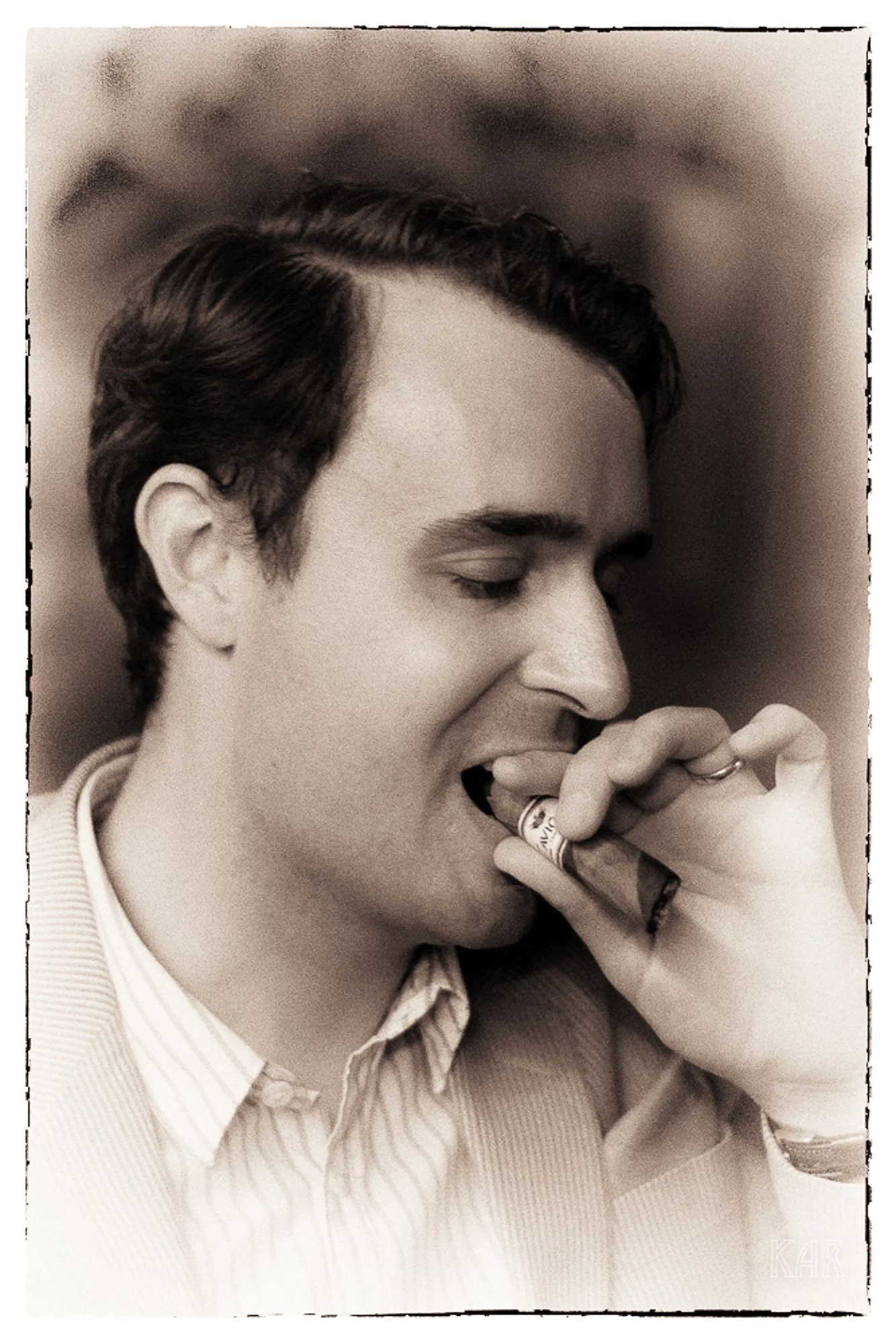 A young man with dark, wavy hair is holding a cigarette near his lips with his eyes closed.