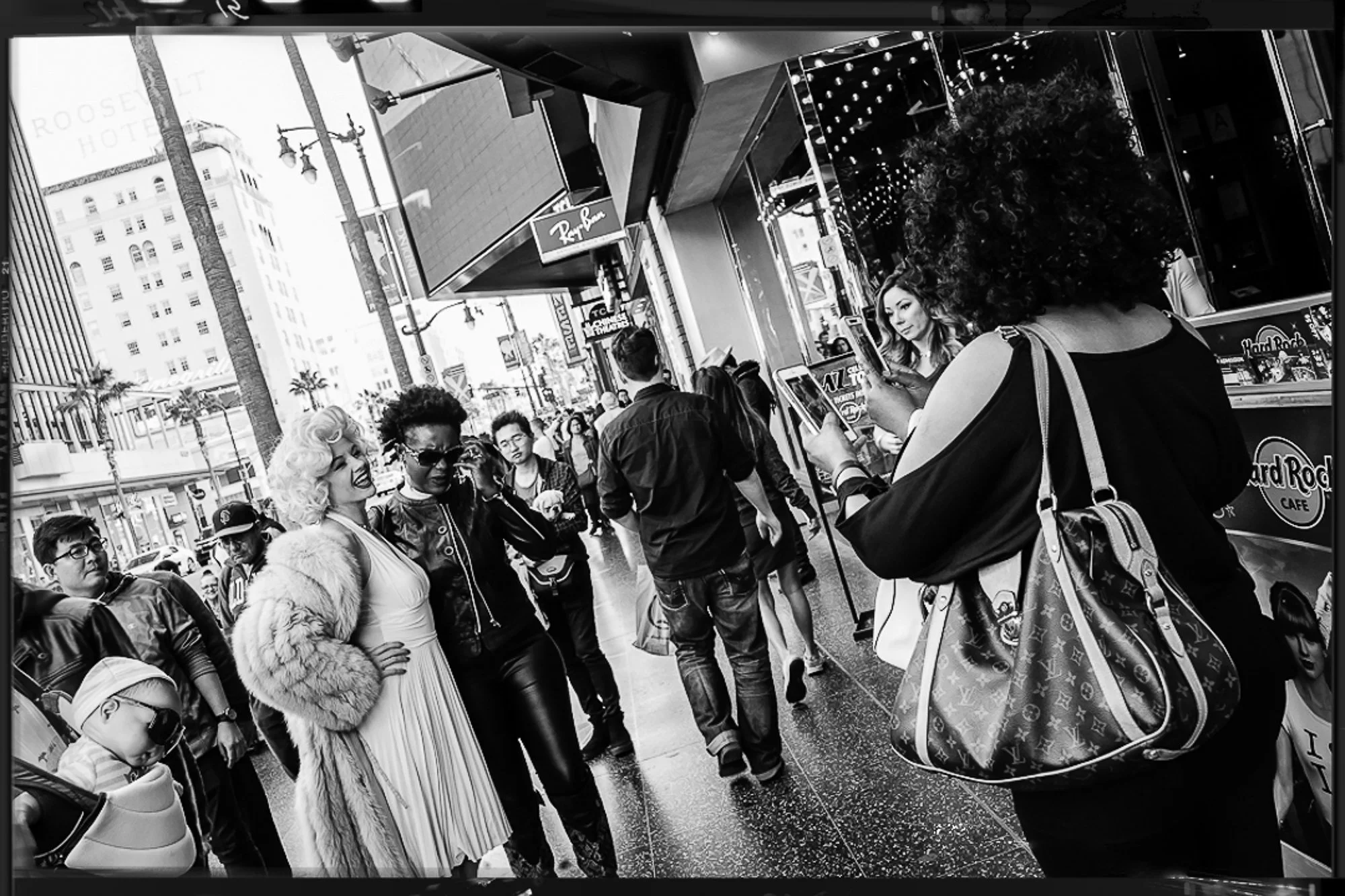 People standing and walking on a busy city sidewalk with storefronts, including a Hard Rock Cafe, and tall buildings in the background.