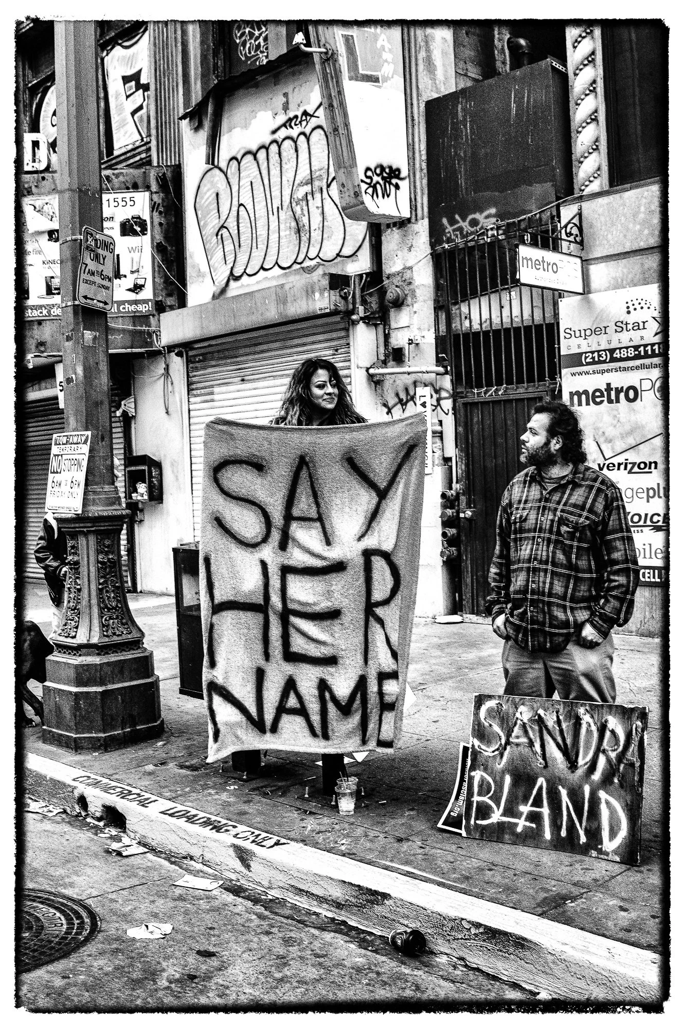 A woman holding a protest sign with the words "SAY HER NAME" written on it, standing on a city sidewalk next to a man. A smaller sign on the ground reads "SANDRA BAND." Graffiti and street signs are visible in the background.