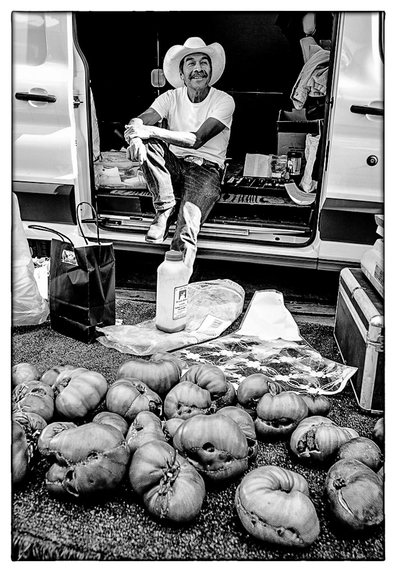 A man wearing a cowboy hat, sitting inside a van with the doors open, surrounded by pumpkins on the ground, with various items and bags inside and outside the van.