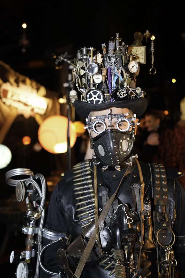 Person wearing steampunk costume with goggles, mask, and a tall hat decorated with gears, clocks, and machinery, standing in a dimly lit environment.