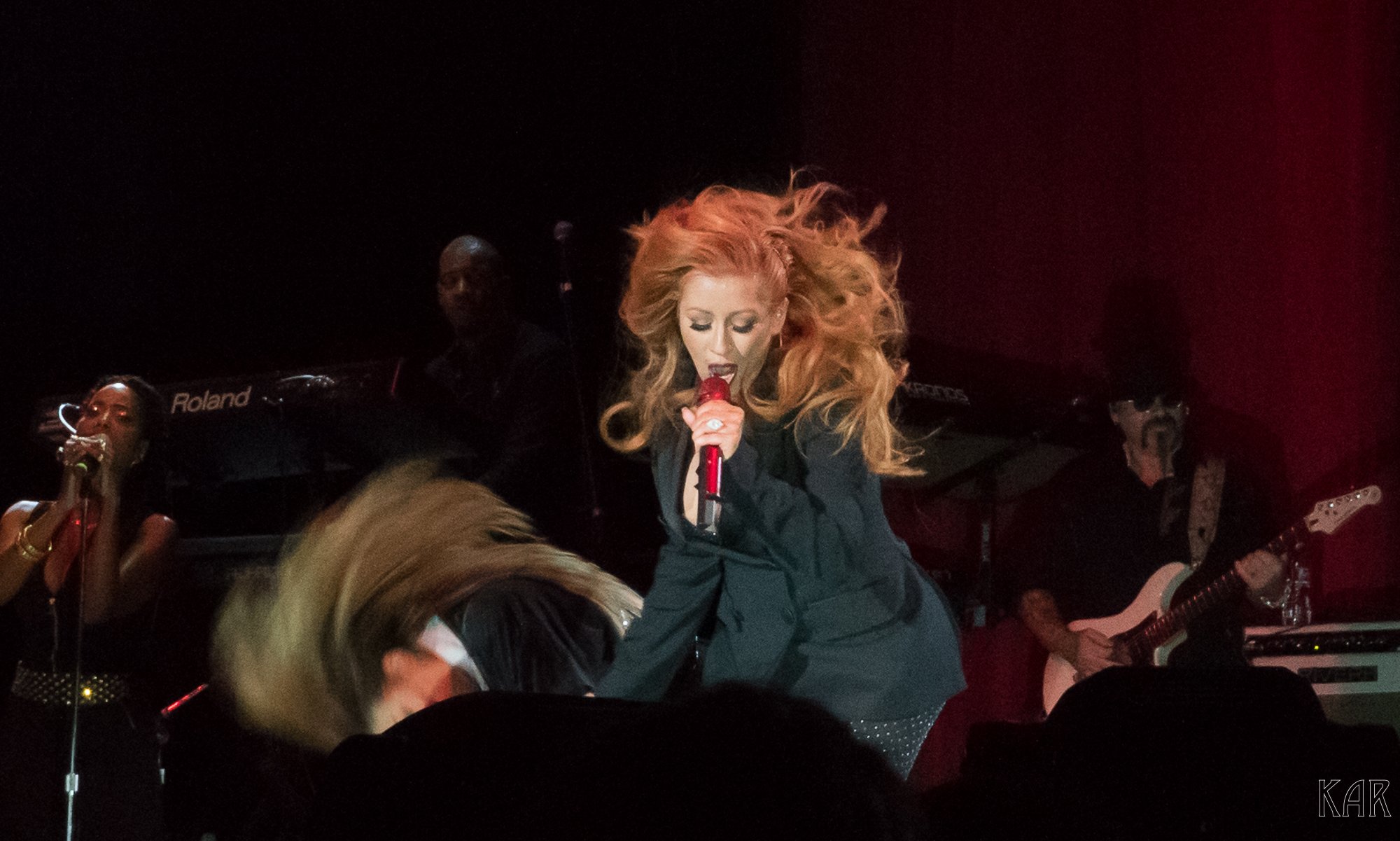 A singer with long, wavy red hair performs on stage, holding a red microphone, with band members playing keyboards and guitar in the background.