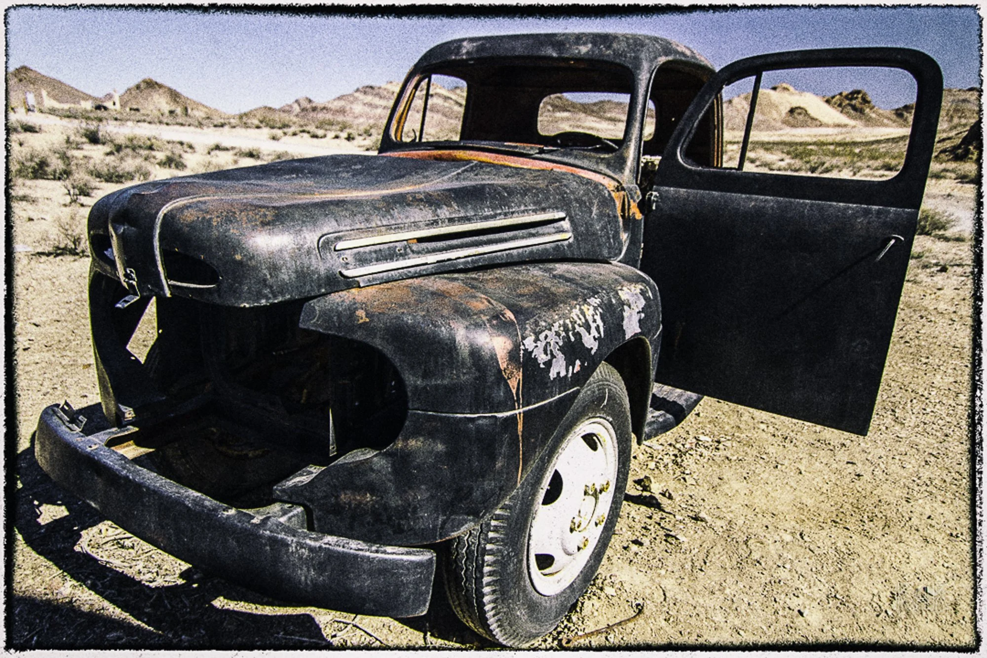An old, rusty, abandoned car in a desert landscape with mountains and sparse vegetation in the background.