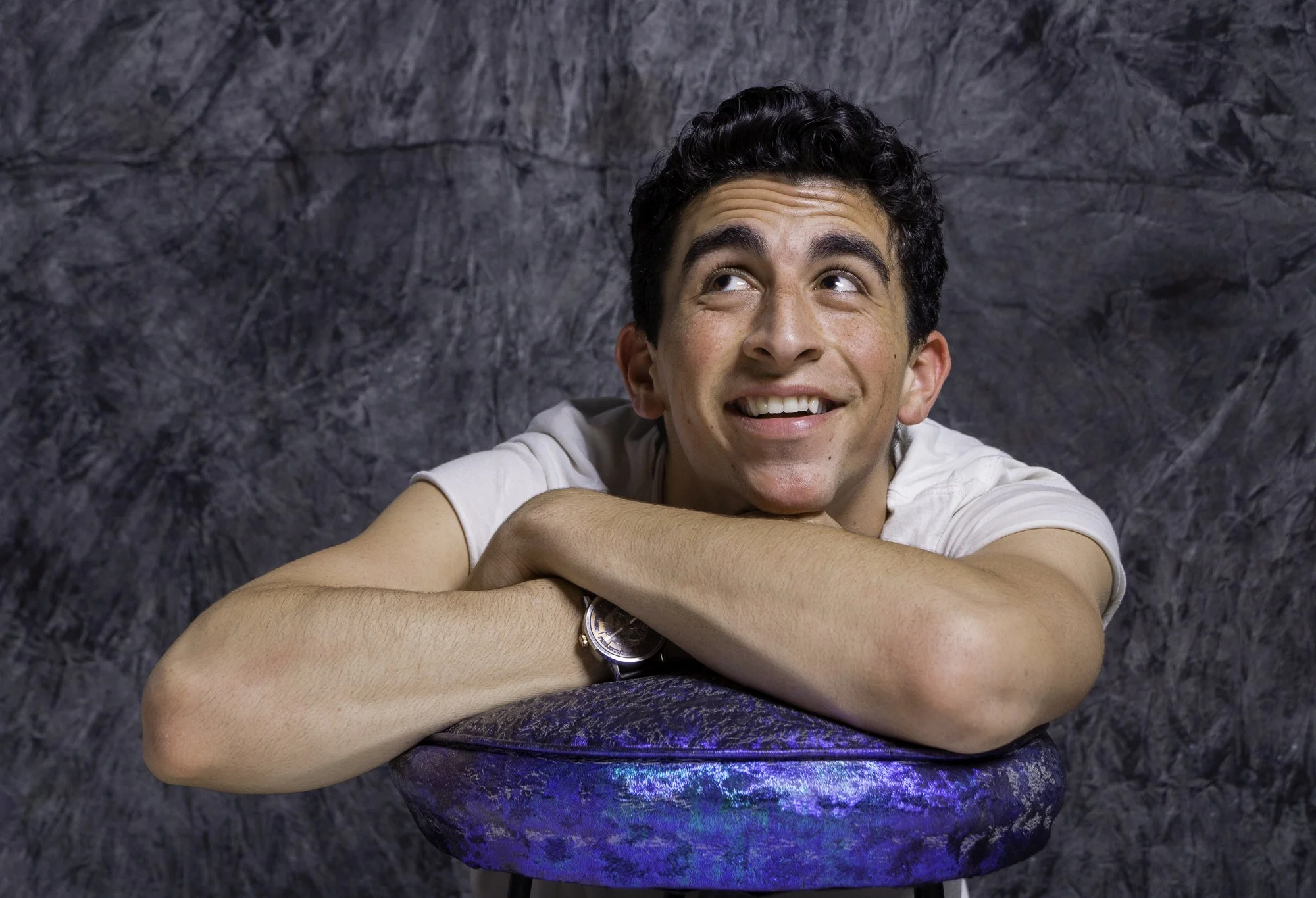 Portrait of a young man with dark curly hair resting his head on his crossed arms on a barstool, smiling and looking up against a gray textured background.