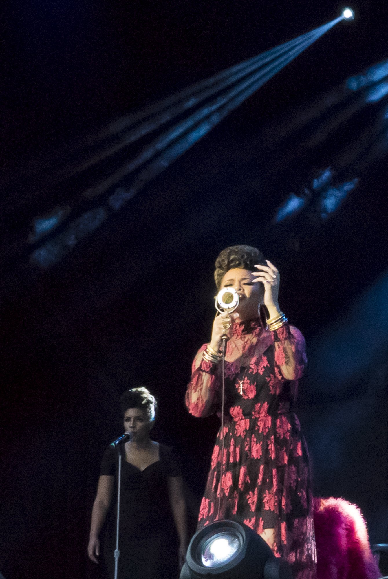 Andra Day singing into a vintage microphone on stage, dressed in a long red and black floral dress, with another woman standing in the background near a microphone, dim stage lighting, and blue spotlights overhead.