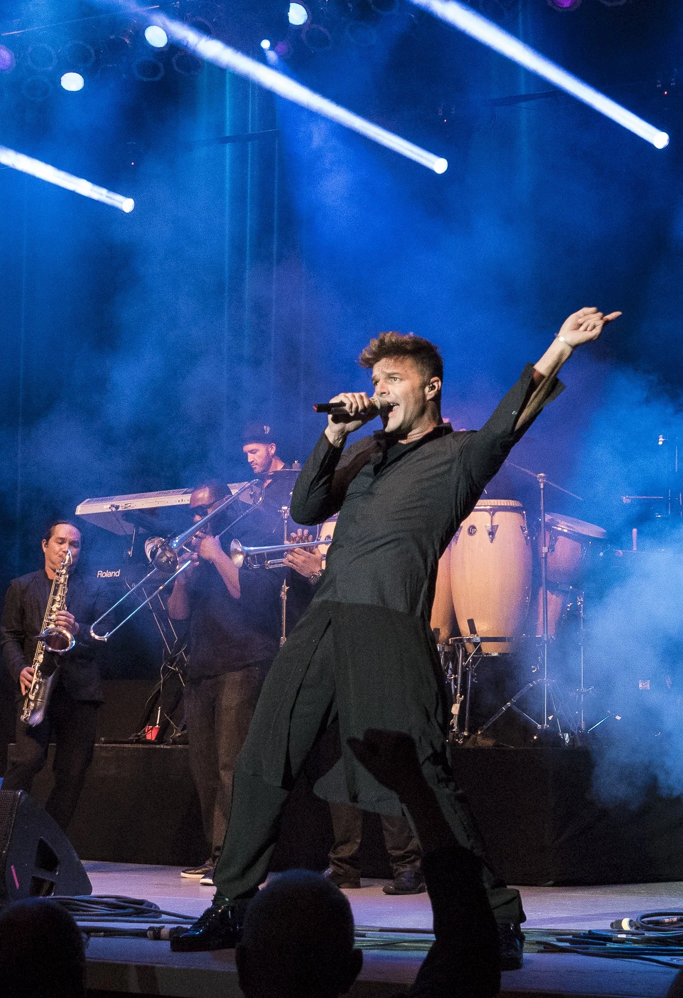Performer singing into a microphone on stage with band members playing musical instruments in the background, blue lighting, and smoke effects.