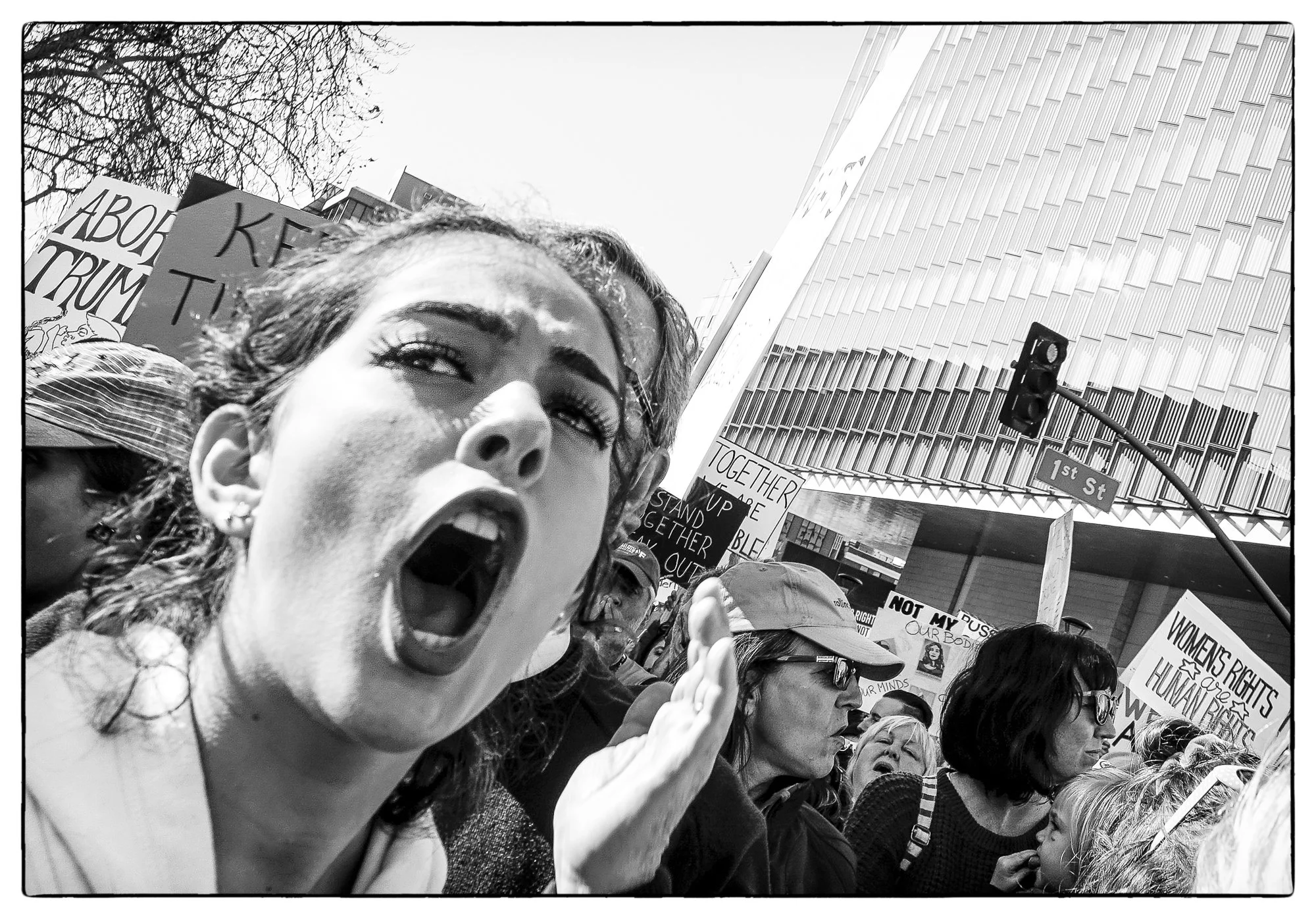 A black and white photo of a woman protesting, shouting passionately with others holding signs. The signs include messages about women's rights and equality, and the protest appears to be on a city street intersection.