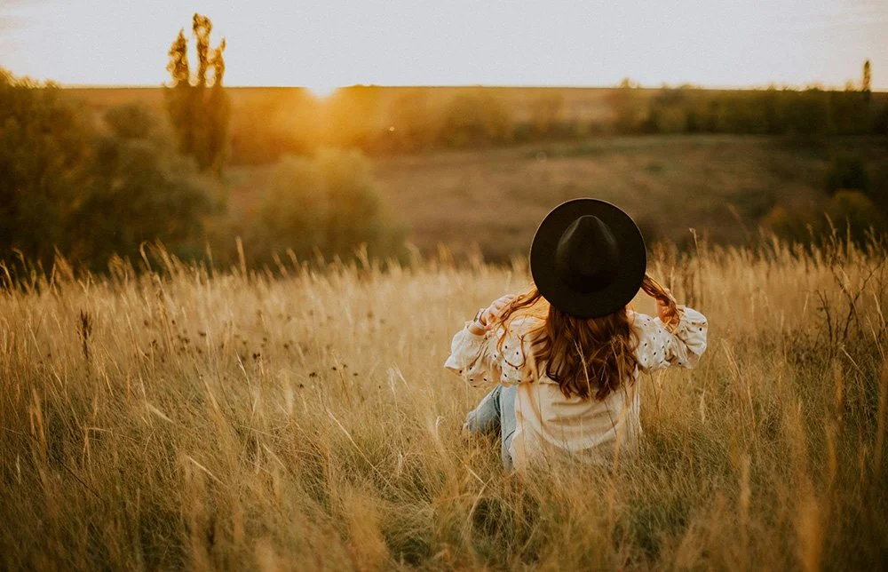 A woman sitting in a grassy field facing away looking at the sunset.