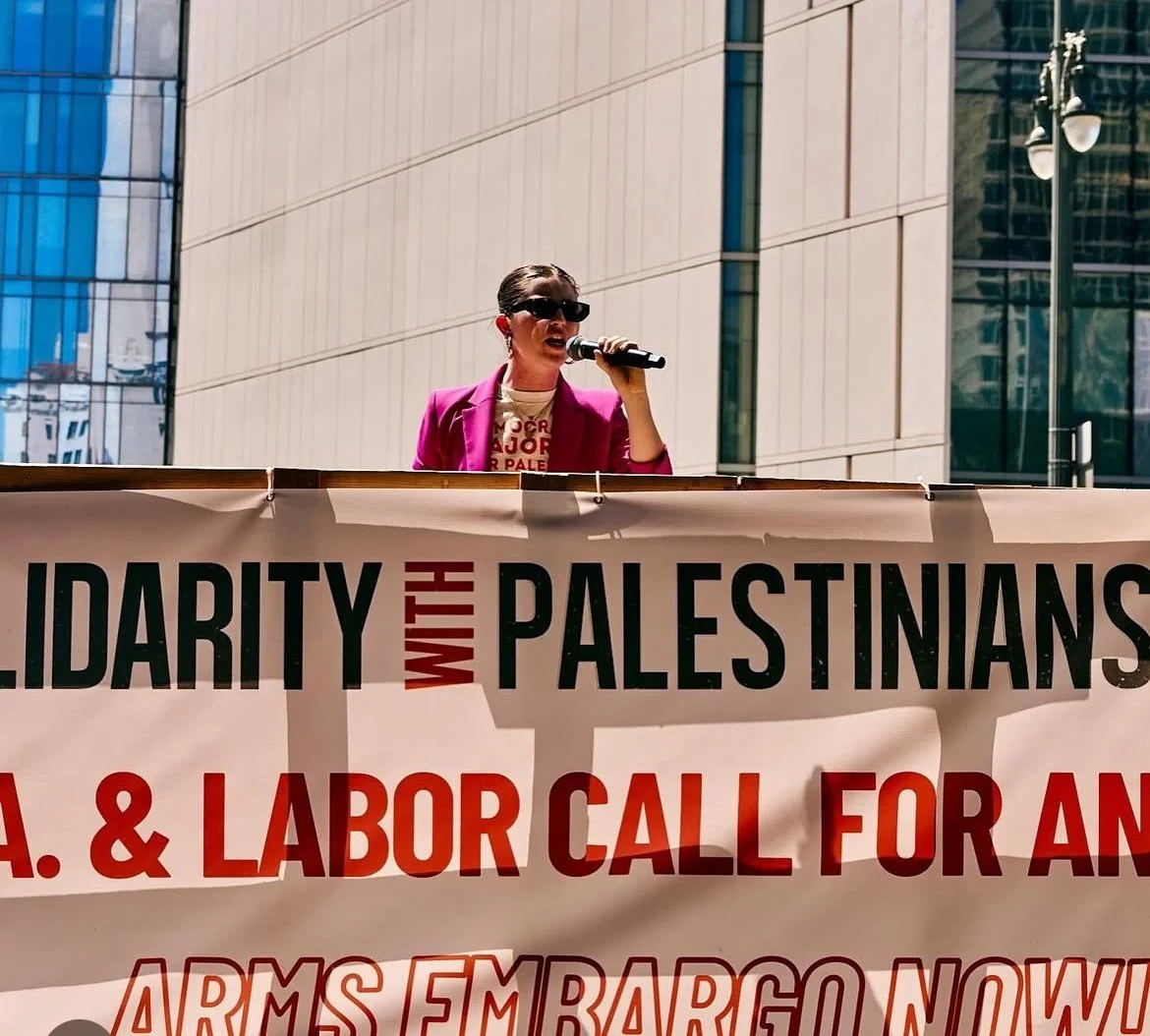 A woman in a pink blazer and sunglasses speaking into a microphone at a protest rally, standing behind a banner in an urban setting with modern buildings in the background.