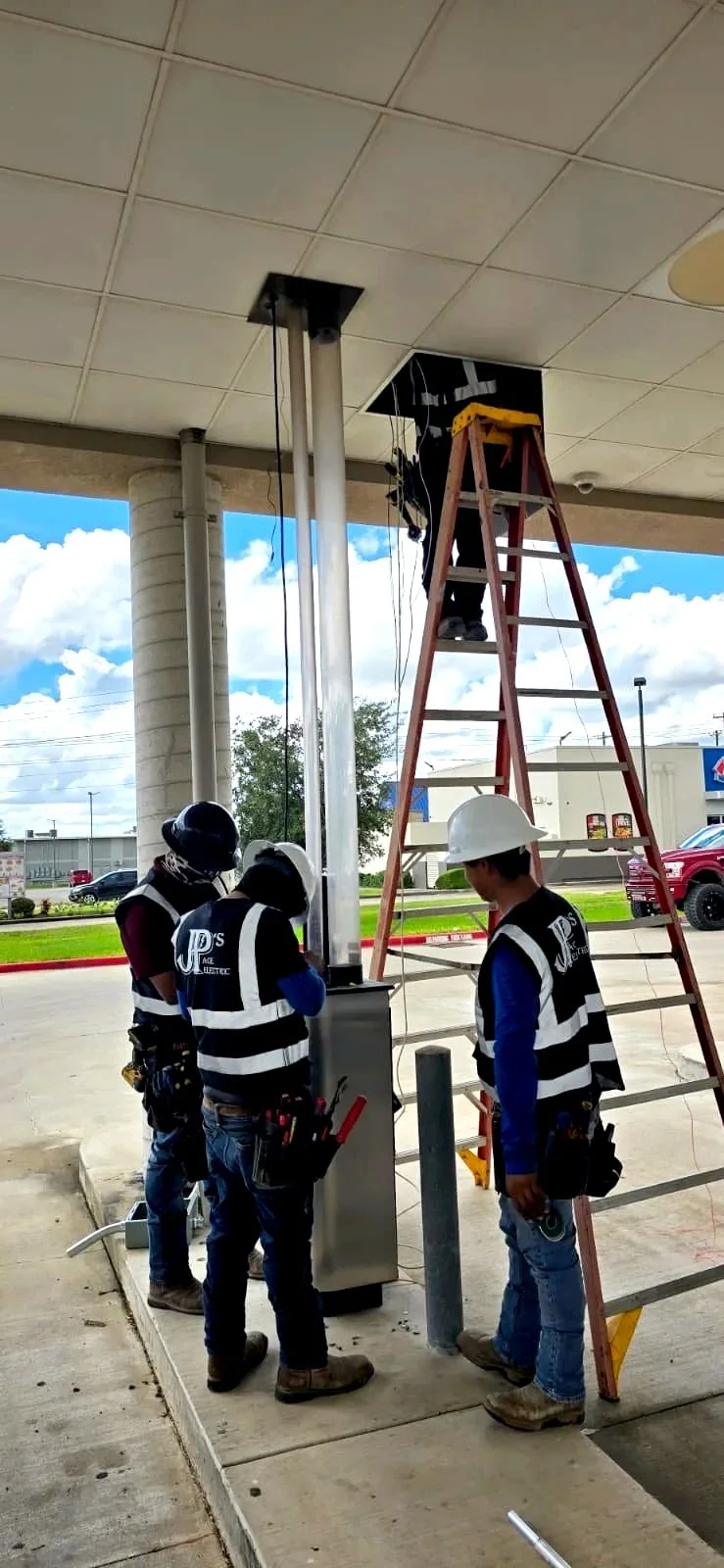 Four construction workers installing electrical equipment outside a building, with one worker on a ladder working on the ceiling and three workers on the ground observing, all wearing safety gear including helmets and vests.