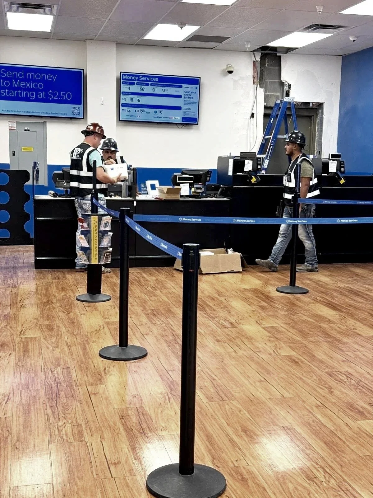 Interior of a financial service counter with two workers wearing safety helmets and vests, and a customer with a helmet. Blue barriers create a queue area, digital screens display money services.