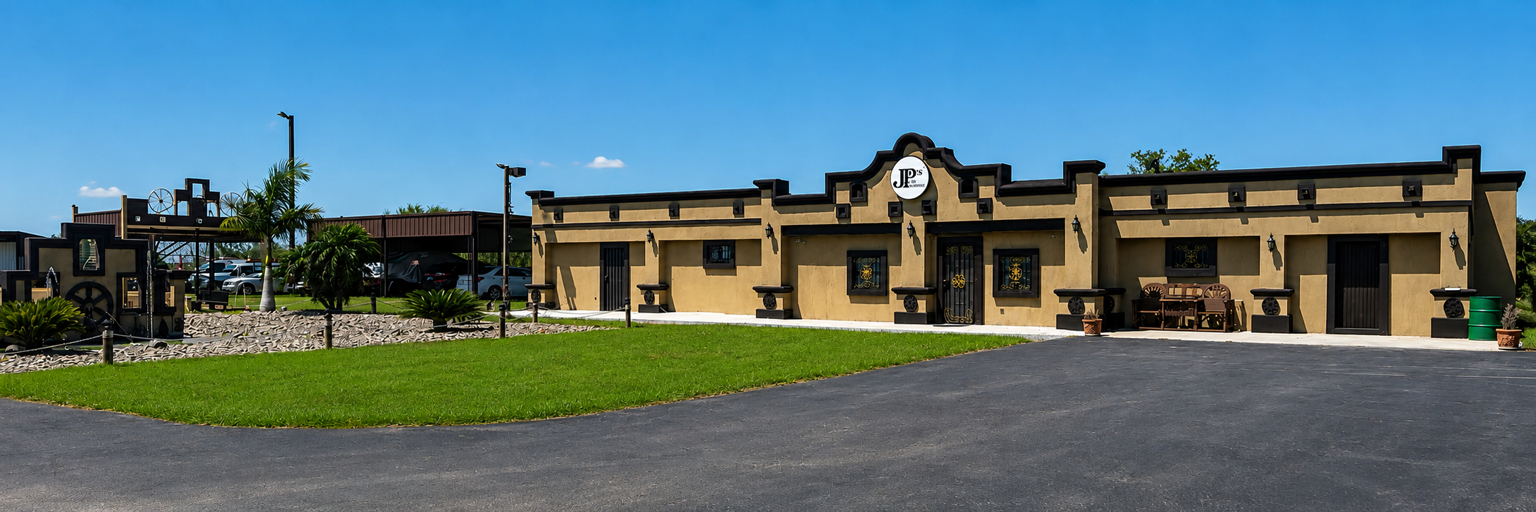 Exterior of a building with a beige facade, black trim, and a sign that reads 'JP's'. The building is decorated in a southwestern style and has small windows with decorative ironwork. There is a parking lot with several parked cars, and a landscaped area with palm trees, bushes, and rocks in the foreground. A bench and potted plants are placed outside the building under a clear blue sky.